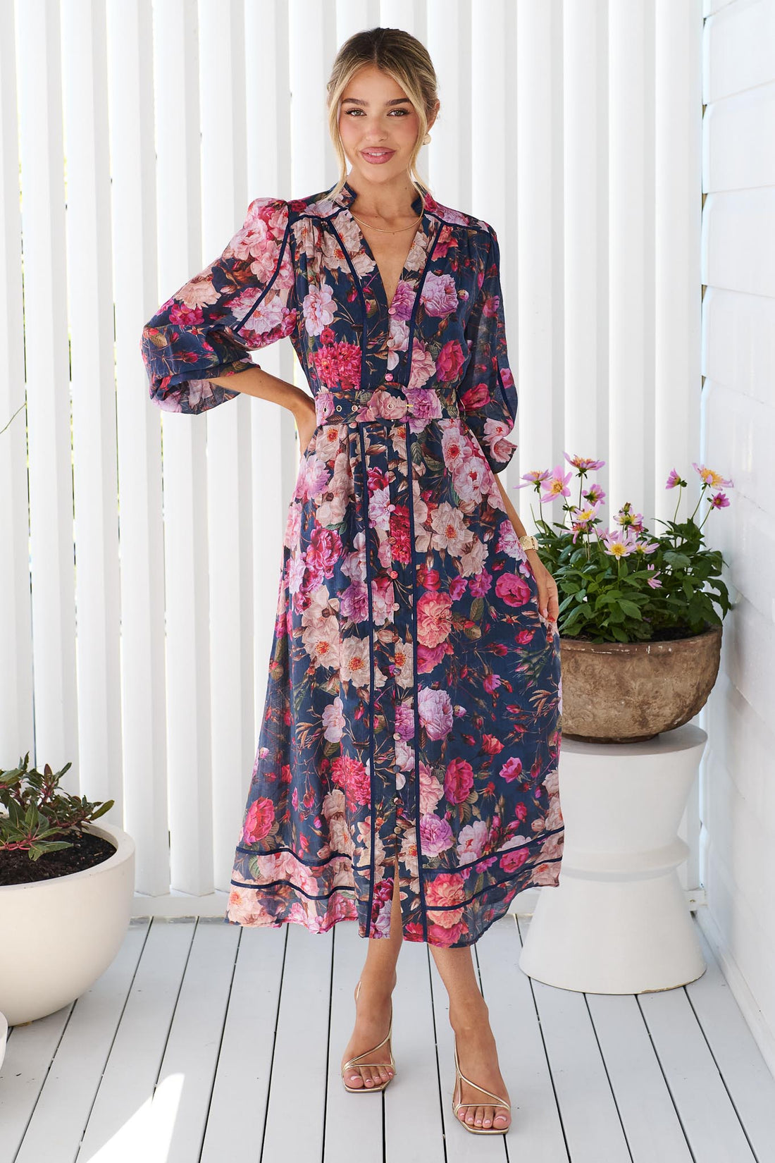 A woman wearing the minnie + MAXXIE Ciara navy blue midi shirt dress with a pink, red, and white floral print, standing on a white deck with potted plants in the background.