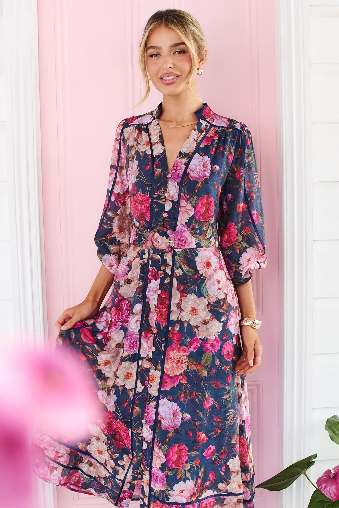 A woman wearing the minnie + MAXXIE Ciara navy blue floral midi dress with pink and red flowers, standing in front of a pink door.