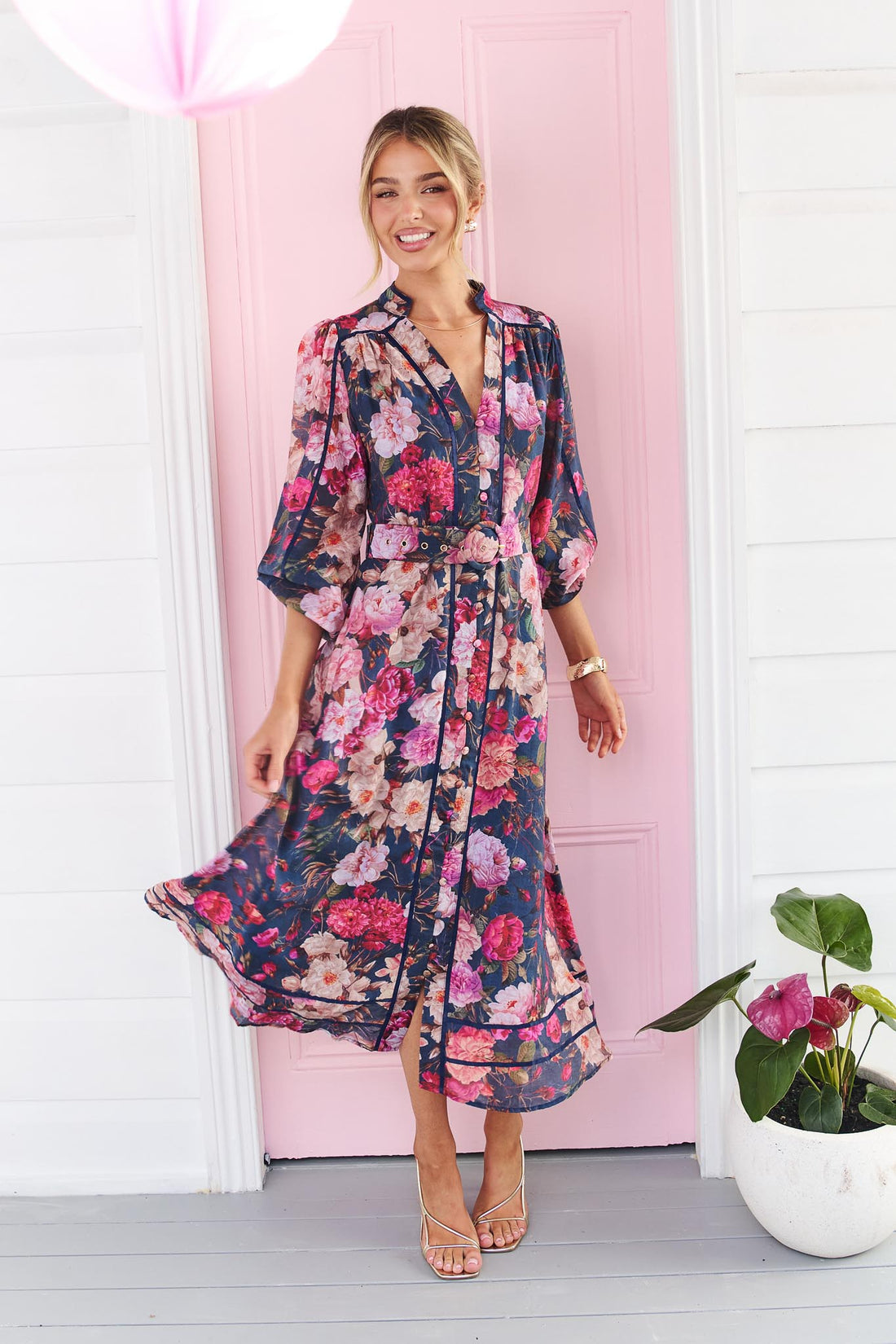 A woman wearing the minnie + MAXXIE Ciara navy blue floral midi dress with pink and red flowers, standing in front of a pink door.