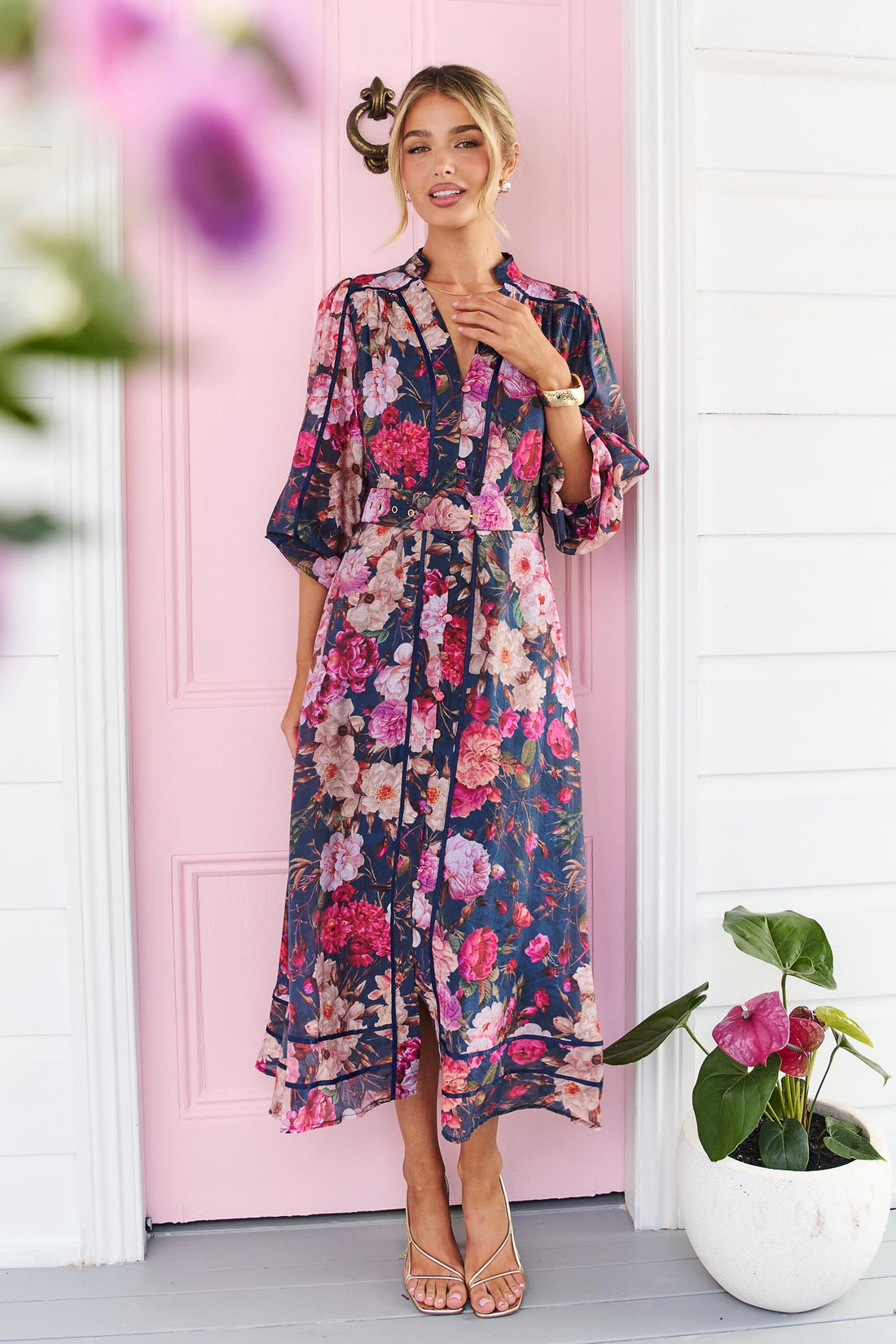 A woman wearing the minnie + MAXXIE Ciara dark navy floral midi dress with pink, red, and white flowers, standing in front of a pink door with a white wall and a potted plant.