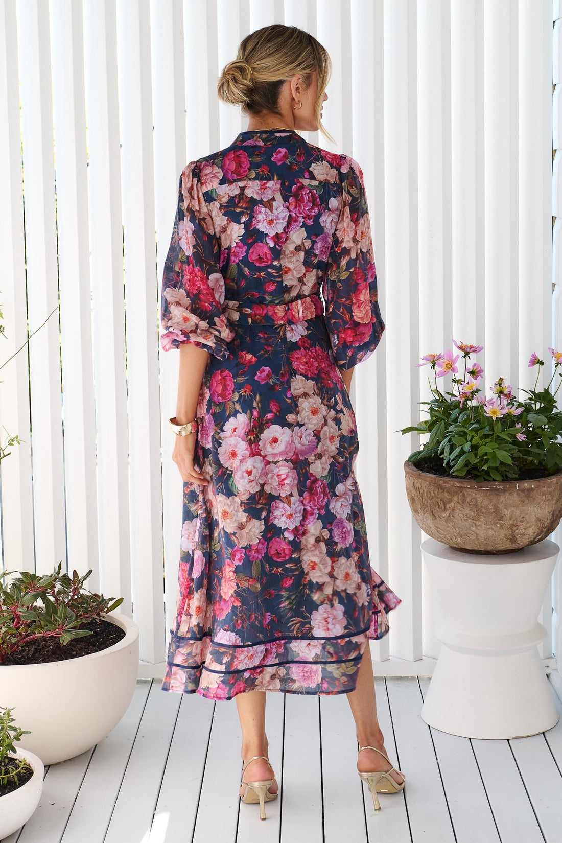 Back view of a woman wearing the minnie + MAXXIE Ciara navy blue midi shirt dress with a pink, red, and white floral print, standing on a white deck with potted plants in the background.