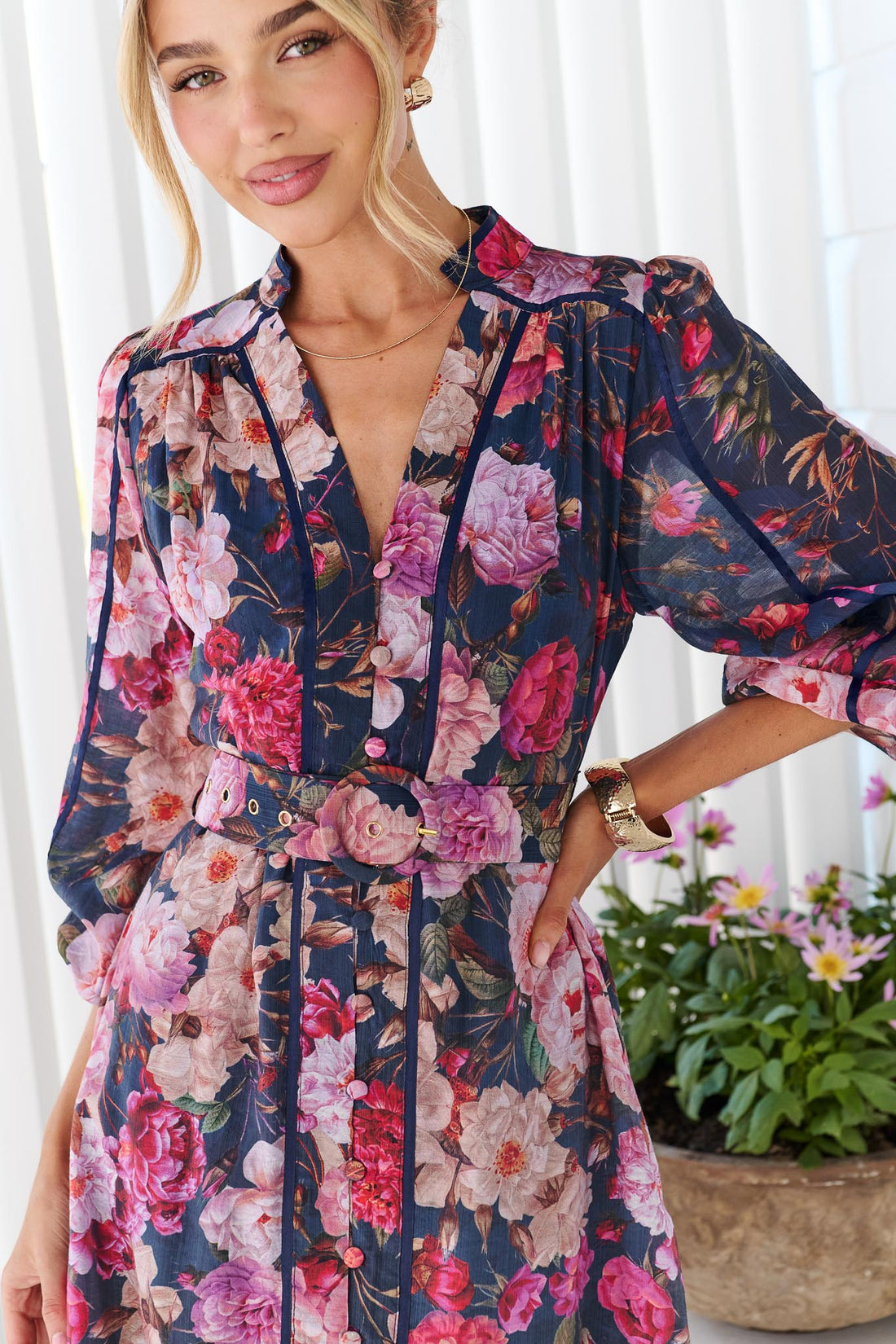 A woman wearing the Ciara navy blue floral print dress with pink, red, and purple flowers, featuring a V-neckline, button front, and matching waist tie belt.