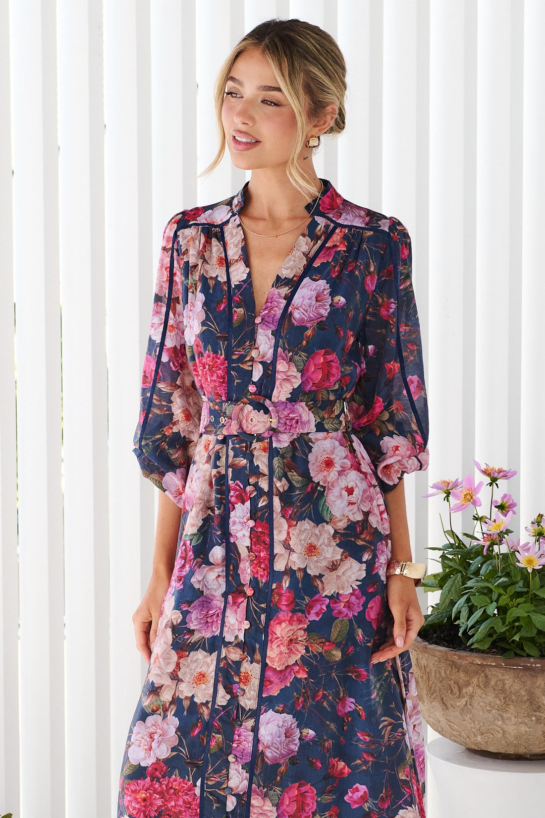 Close up of a woman wearing the minnie + MAXXIE Ciara navy blue midi shirt dress with a pink, red, and white floral print, standing on a white deck with potted plants in the background.