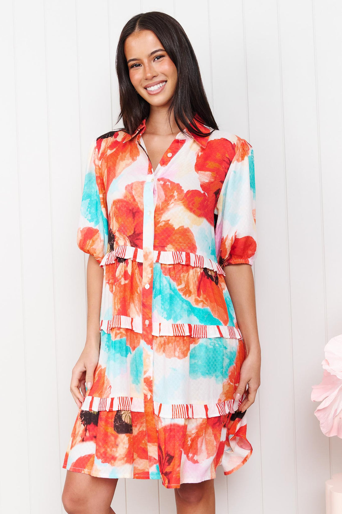 A woman wearing the minnie + MAXXIE short-sleeve Clancy Tiered Dress with a vibrant watercolour floral print in orange, red, turquoise and white, standing in front of a white panelled wall.