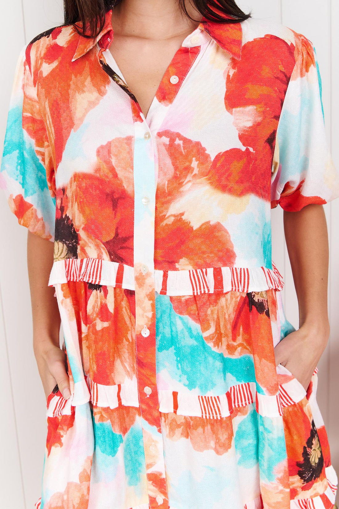 Detail of a woman wearing the minnie + MAXXIE short-sleeve Clancy Tiered Dress with a vibrant watercolour floral print in orange, red, turquoise and white.
