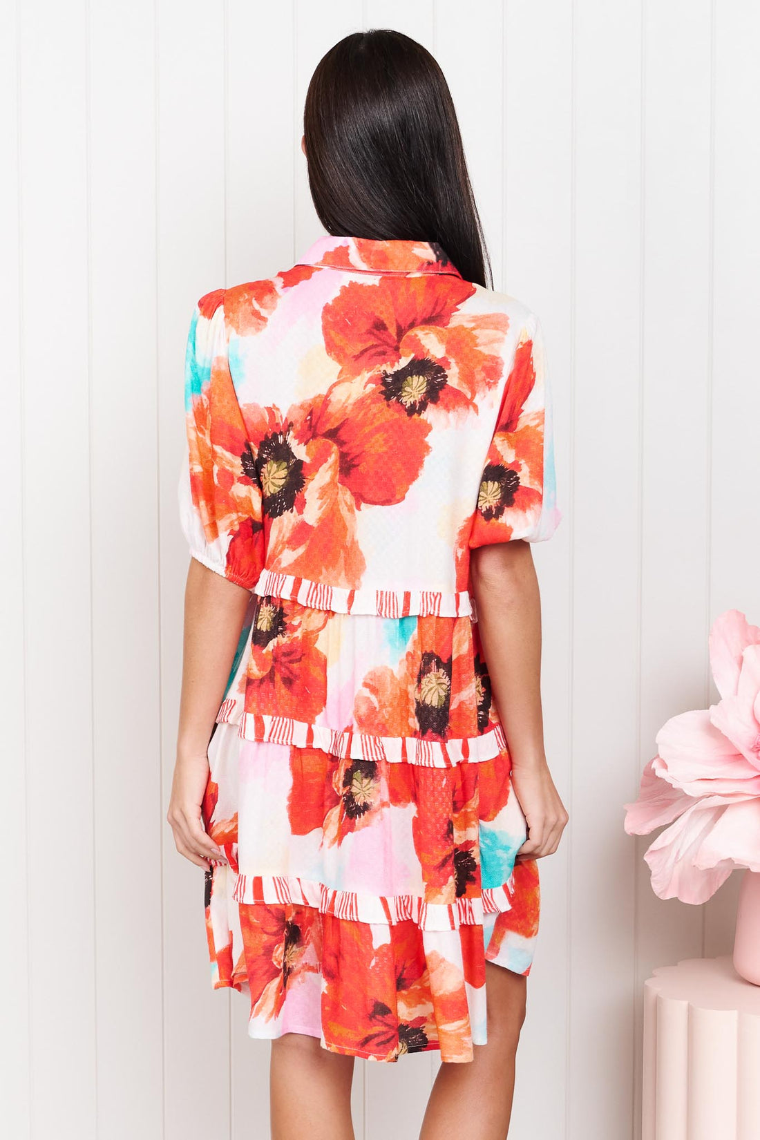 Back view of a woman wearing the minnie + MAXXIE short-sleeve Clancy Tiered Dress with a vibrant watercolour floral print in orange, red, turquoise and white, standing in front of a white panelled wall.