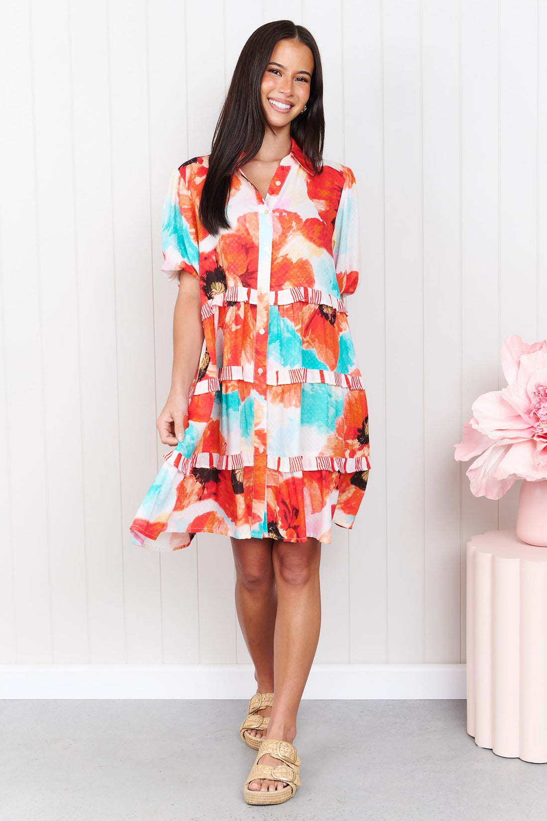 A woman wearing the minnie + MAXXIE short-sleeve Clancy Tiered Dress with a vibrant watercolour floral print in orange, red, turquoise and white, standing in front of a white panelled wall.