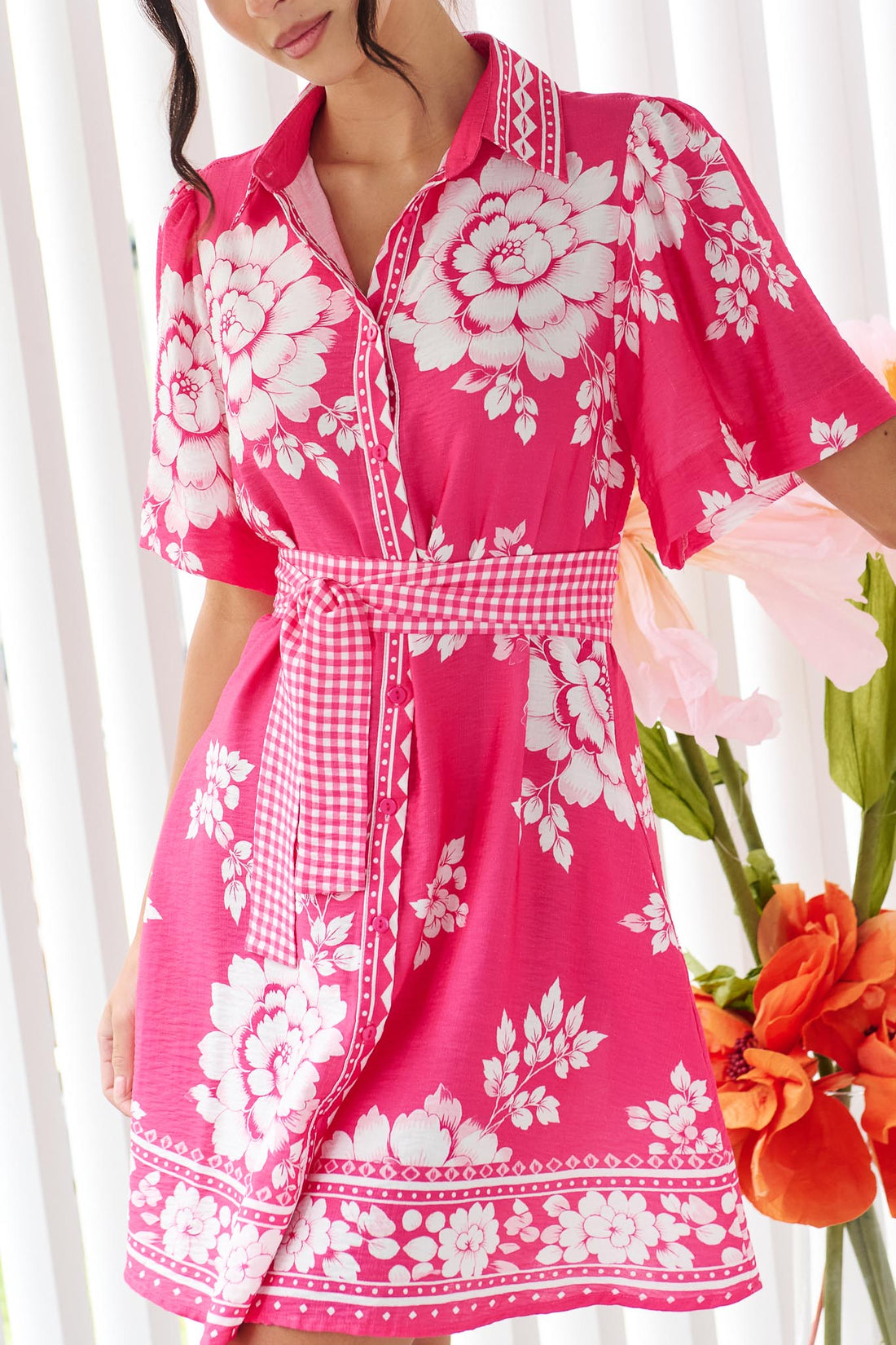 Detail of a woman wearing the minnie + MAXXIE Cleo dress in a hot pink and white floral print with short-sleeves and a matching gingham waist tie.