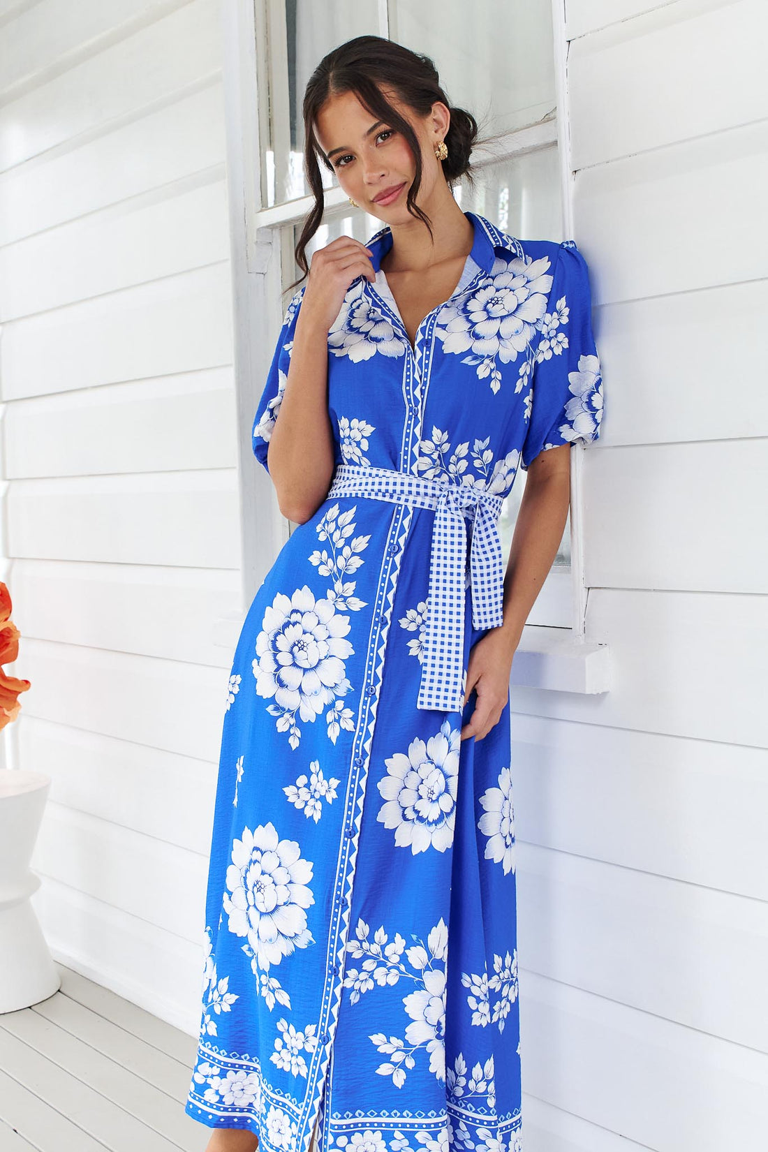 A woman wearing the minnie + MAXXIE Cleo Midi Dress in a bright cobalt blue and white floral print with a gingham waist tie, standing in front of a white panelled wall.