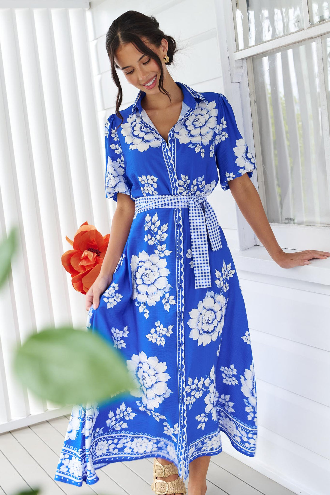 A woman wearing the minnie + MAXXIE Cleo Midi Dress in a bright cobalt blue and white floral print with a gingham waist tie, standing in front of a white panelled wall.