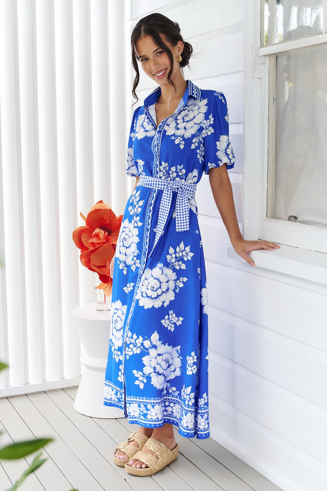 A woman wearing the minnie + MAXXIE Cleo Midi Dress in a bright cobalt blue and white floral print with a gingham waist tie, standing in front of a white panelled wall.