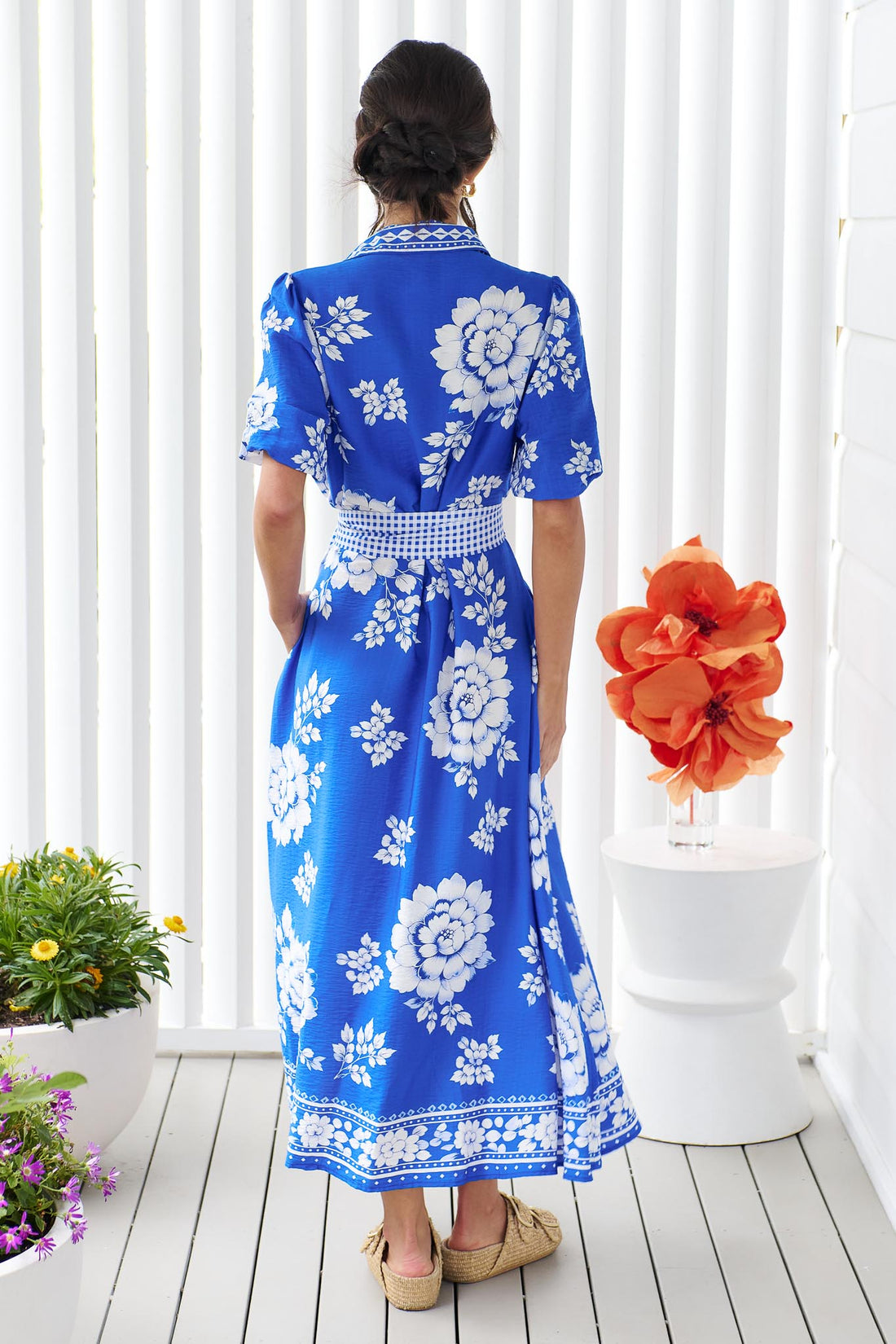 Back view of a woman wearing the minnie + MAXXIE Cleo Midi Dress in a bright cobalt blue and white floral print with a gingham waist tie, standing in front of a white panelled wall.