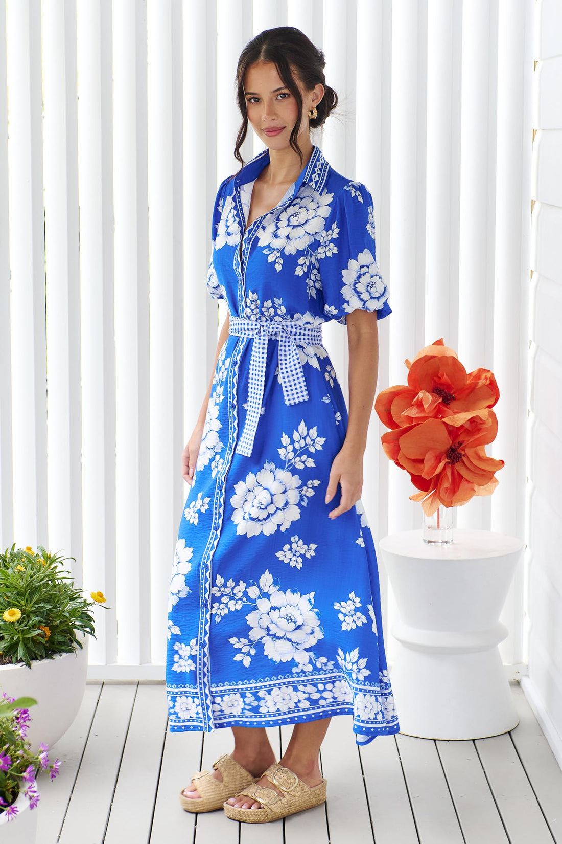 A woman wearing the minnie + MAXXIE Cleo Midi Dress in a bright cobalt blue and white floral print with a gingham waist tie, standing in front of a white panelled wall.
