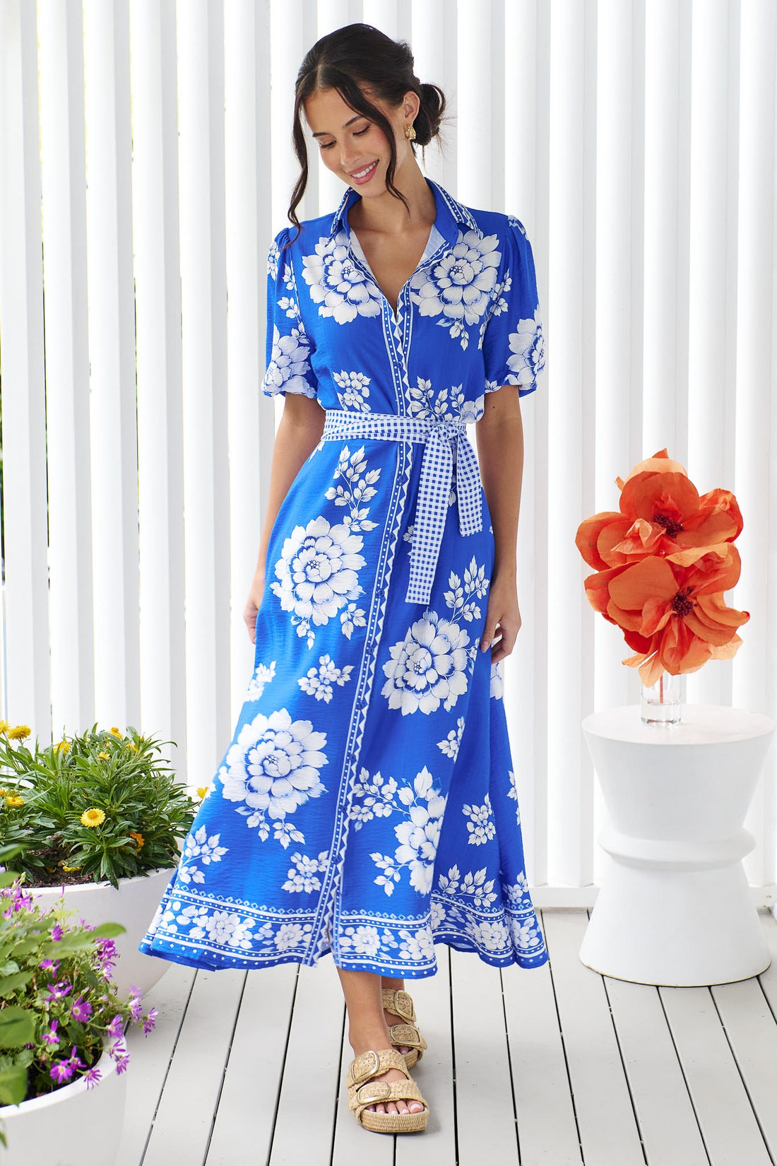 A woman wearing the minnie + MAXXIE Cleo Midi Dress in a bright cobalt blue and white floral print with a gingham waist tie, standing in front of a white panelled wall.