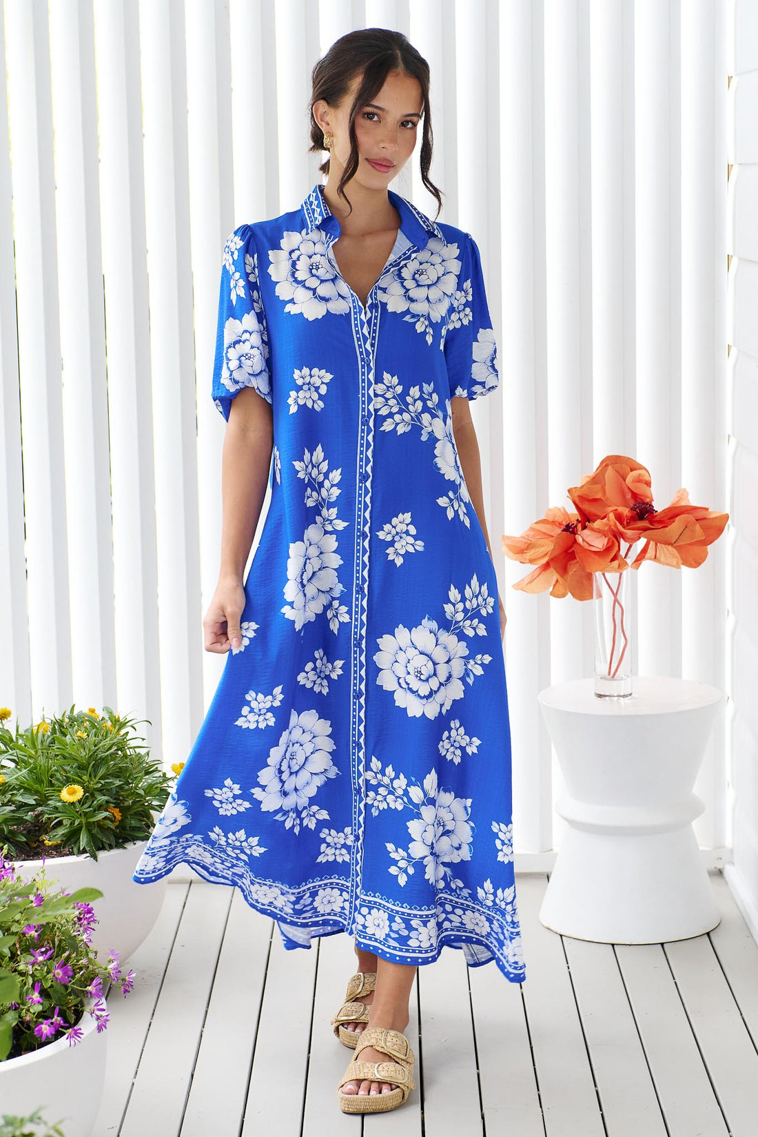 A woman wearing the minnie + MAXXIE Cleo Midi Dress in a bright cobalt blue and white floral print, standing in front of a white panelled wall.