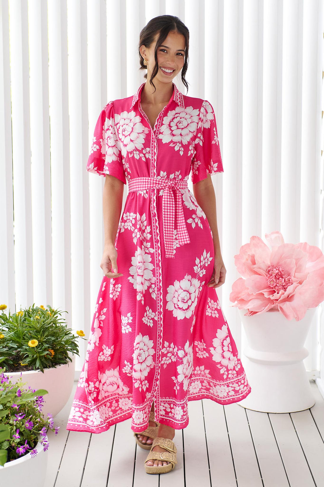 A woman wearing the minnie + MAXXIE Cleo Midi Dress in a hot pink and white floral print with a gingham waist tie, standing in front of a white panelled wall.