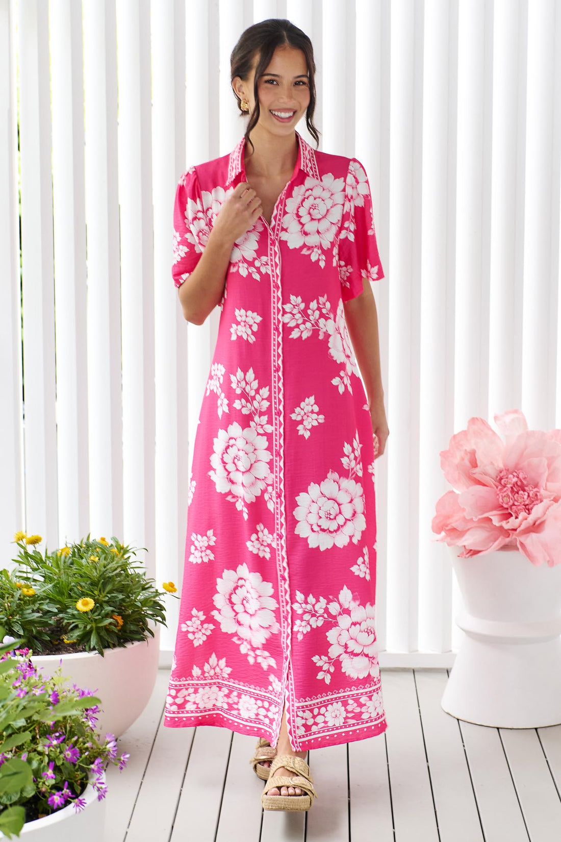 A woman wearing the minnie + MAXXIE Cleo Midi Dress in a hot pink and white floral print, standing in front of a white panelled wall.