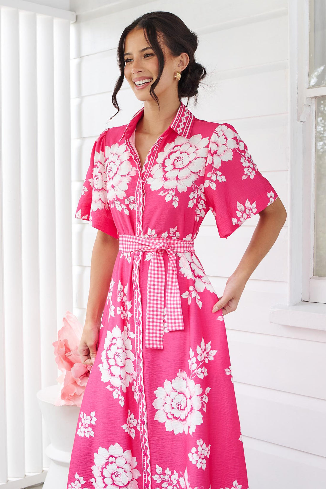 A woman wearing the minnie + MAXXIE Cleo Midi Dress in a hot pink and white floral print with a gingham waist tie, standing in front of a white panelled wall.