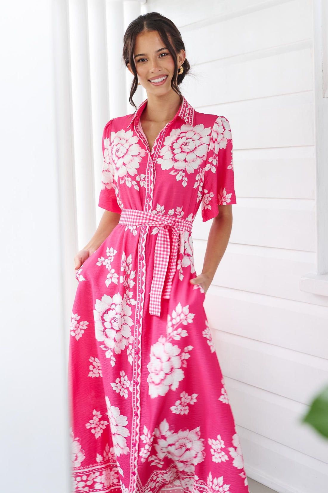 A woman wearing the minnie + MAXXIE Cleo Midi Dress in a hot pink and white floral print with a gingham waist tie, standing in front of a white panelled wall.