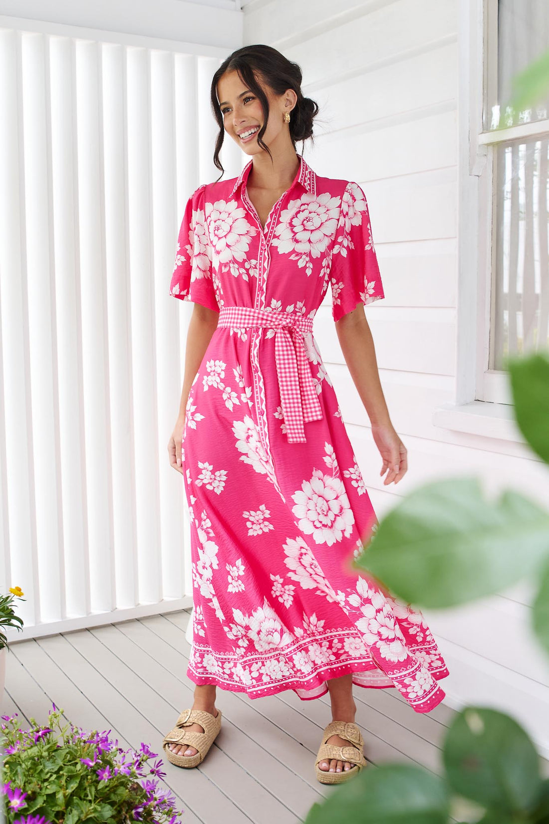 A woman wearing the minnie + MAXXIE Cleo Midi Dress in a hot pink and white floral print with a gingham waist tie, standing in front of a white panelled wall.
