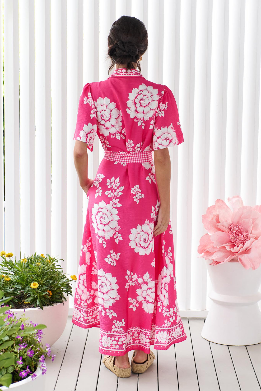 Back view of a woman wearing the minnie + MAXXIE Cleo Midi Dress in a hot pink and white floral print with a gingham waist tie, standing in front of a white panelled wall.