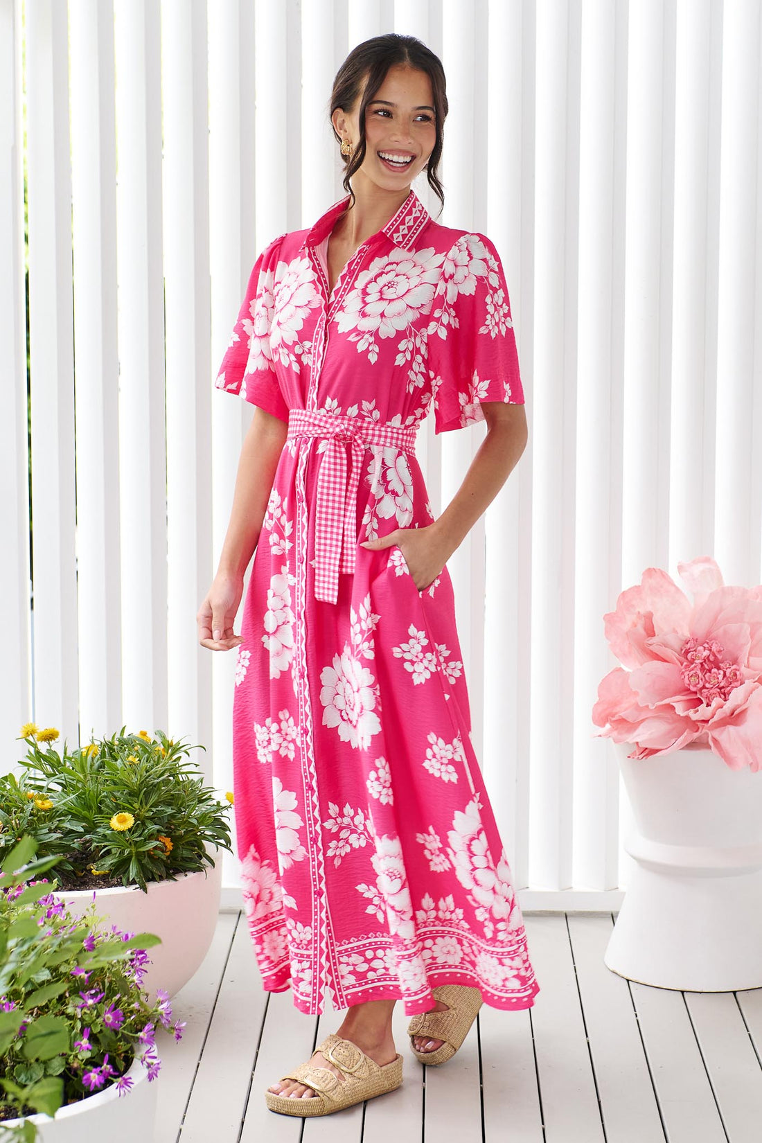 A woman wearing the minnie + MAXXIE Cleo Midi Dress in a hot pink and white floral print with a gingham waist tie, standing in front of a white panelled wall.