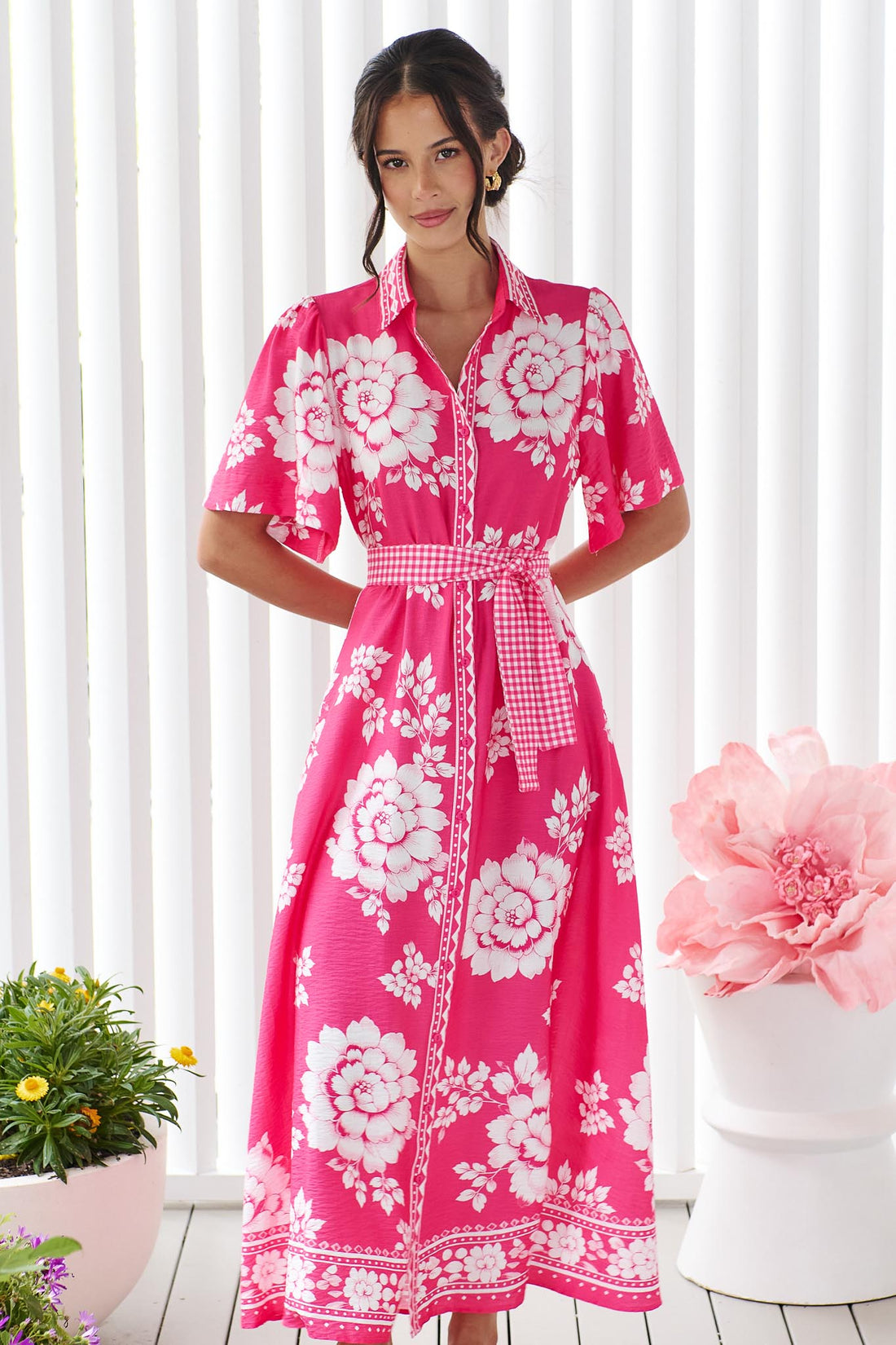 A woman wearing the minnie + MAXXIE Cleo Midi Dress in a hot pink and white floral print with a gingham waist tie, standing in front of a white panelled wall.