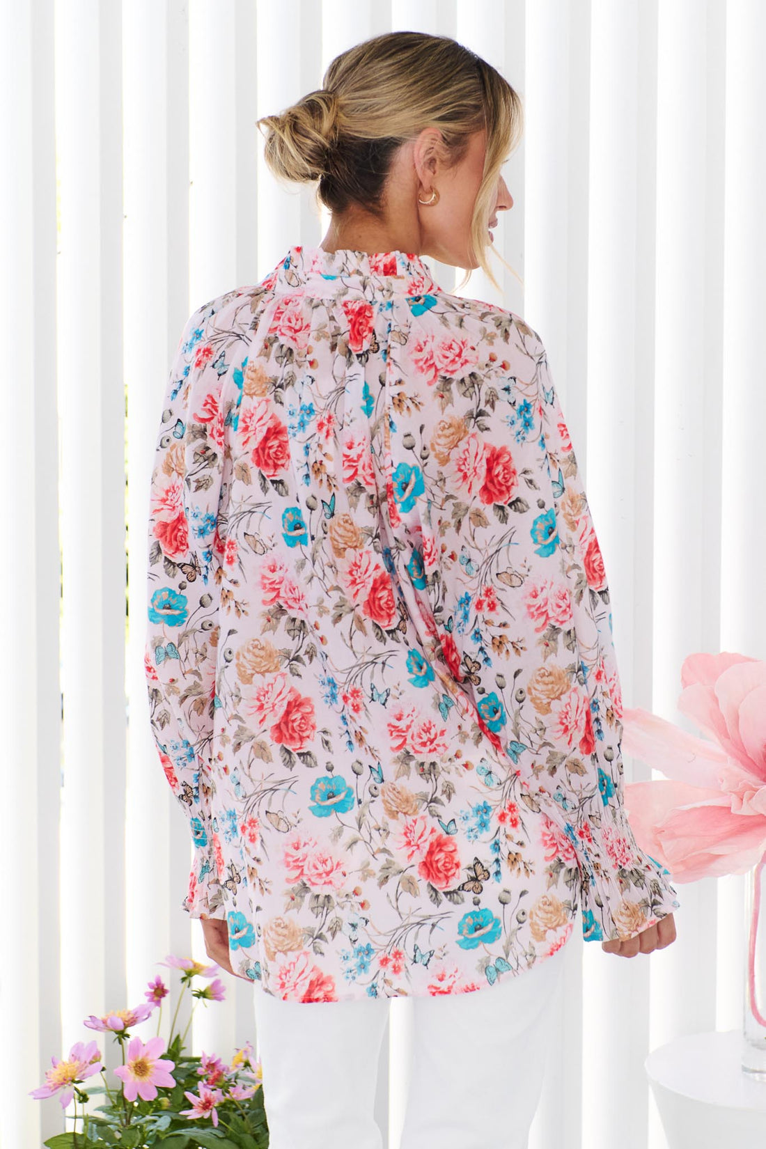 Back view of a woman wearing the minnie + MAXXIE Cleo Floral Shirt, paired with white pants, standing in front of a white vertical panelled wall.