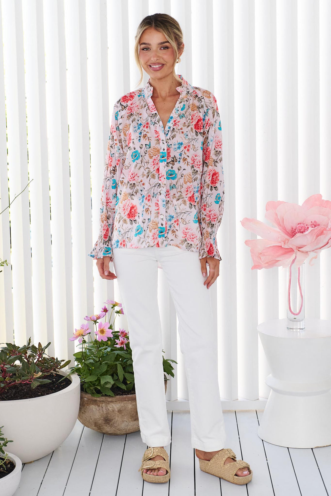A woman wearing the minnie + MAXXIE Cleo Floral Shirt, paired with white pants, standing in front of a white vertical panelled wall with potted flowers.