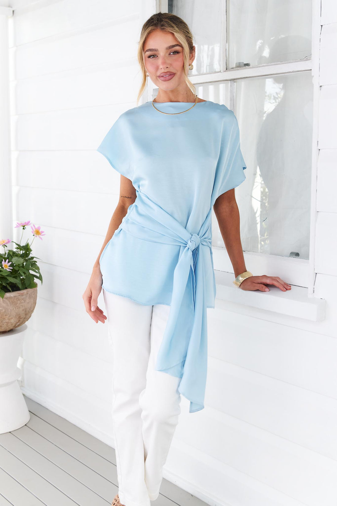 A woman wearing the minnie + MAXXIE Dana satin tie front top in light blue, paired with white jeans, standing in front of a bright white porch setting.