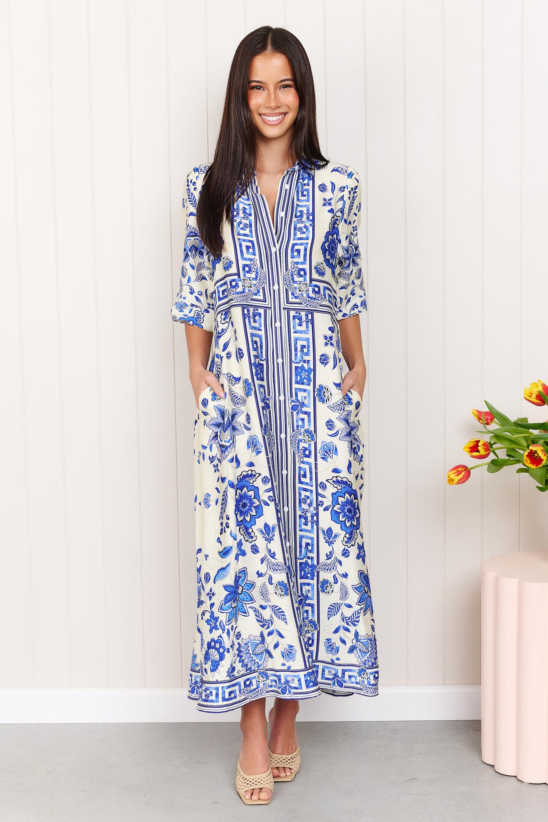 A woman wearing the minnie + MAXXIE Del Mar blue and white floral print midi dress with 3/4 sleeves, standing in front of a white panelled wall.
