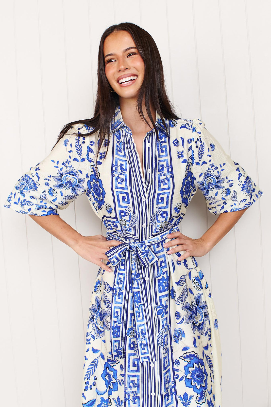 A woman wearing the minnie + MAXXIE Del Mar blue and white floral print midi dress with a tie waist and 3/4 sleeves, standing in front of a white panelled wall.