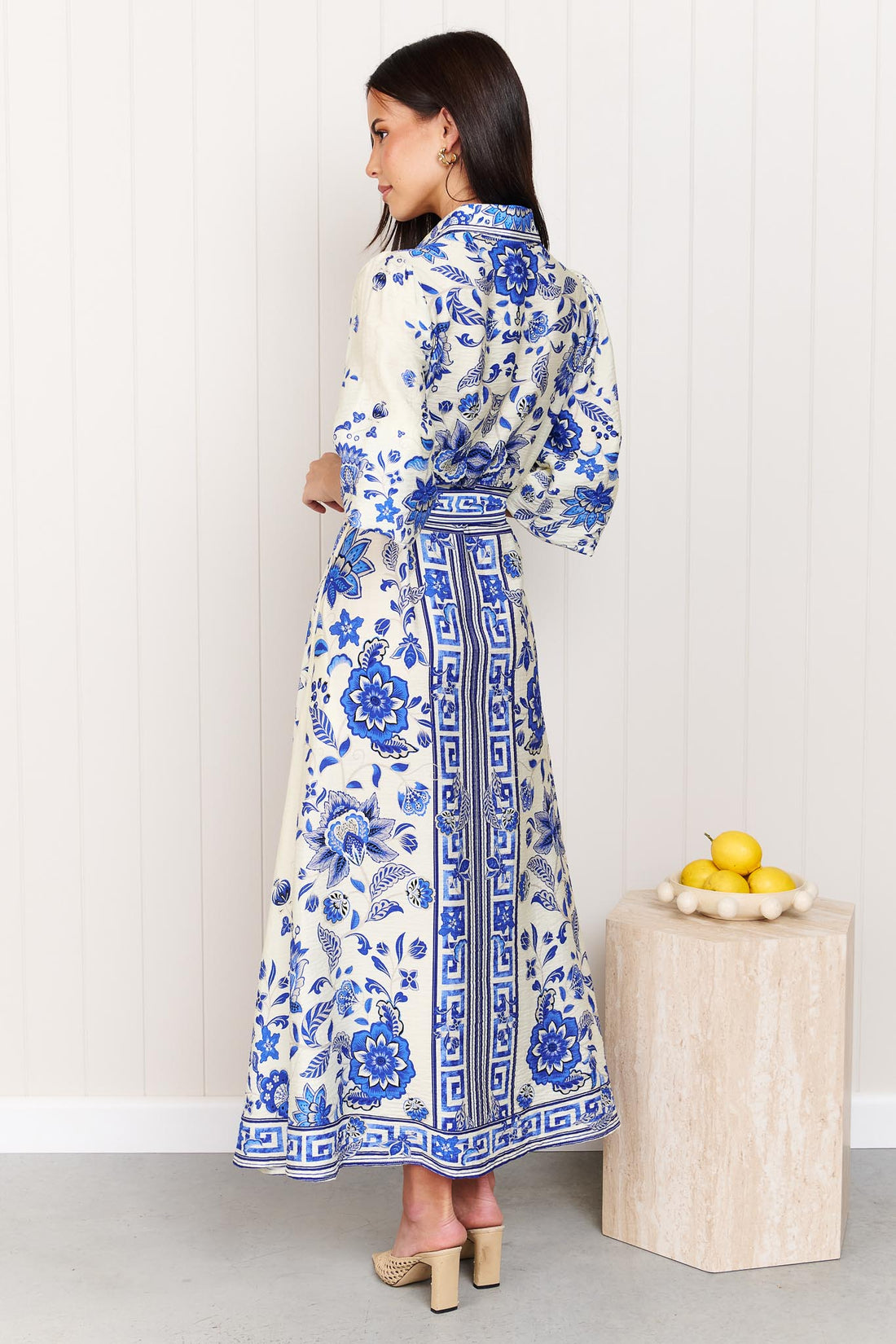 Back view of a woman wearing the minnie + MAXXIE Del Mar blue and white floral print midi dress with a tie waist and 3/4 sleeves, standing in front of a white panelled wall.