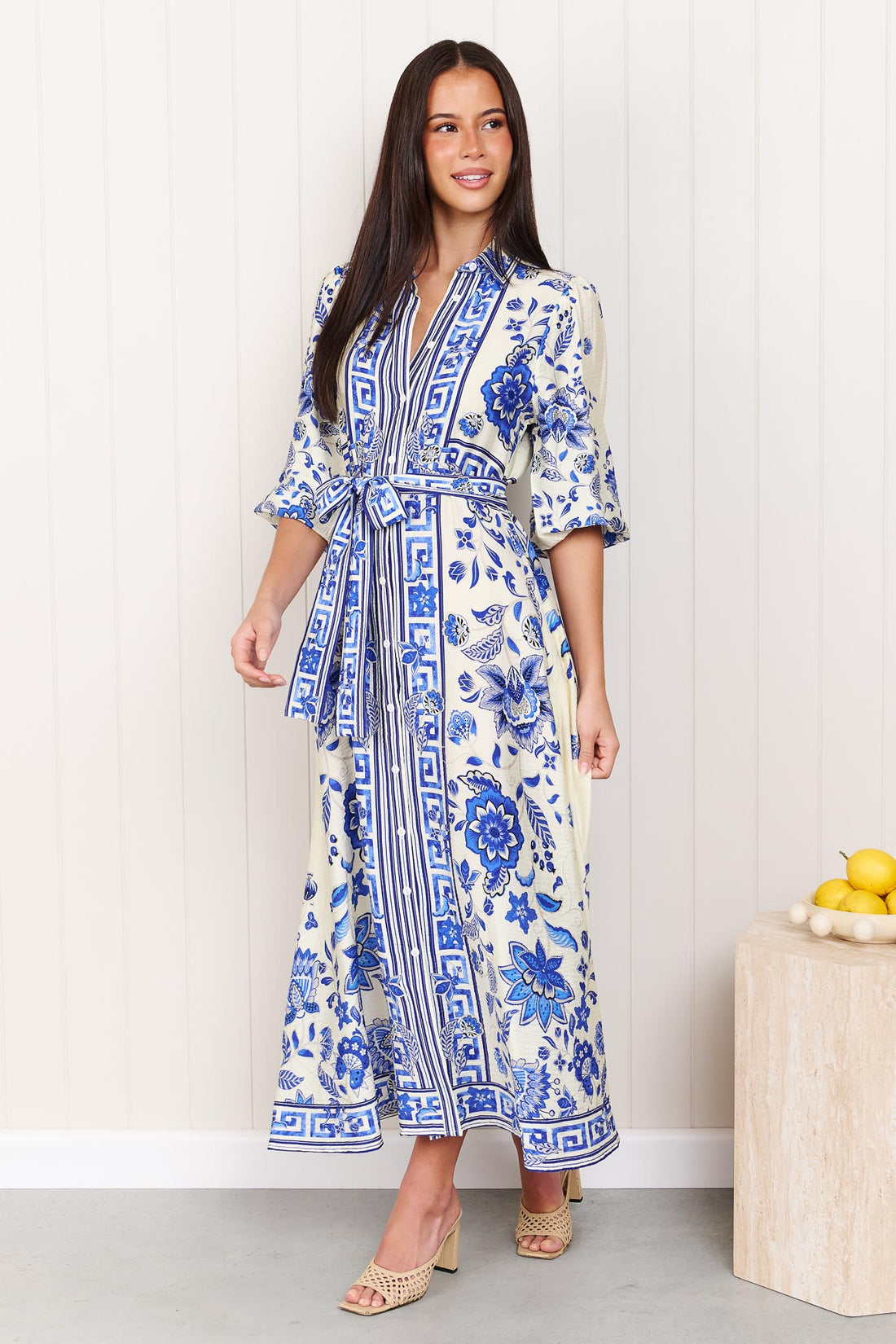 A woman wearing the minnie + MAXXIE Del Mar blue and white floral print midi dress with a tie waist and 3/4 sleeves, standing in front of a white panelled wall.