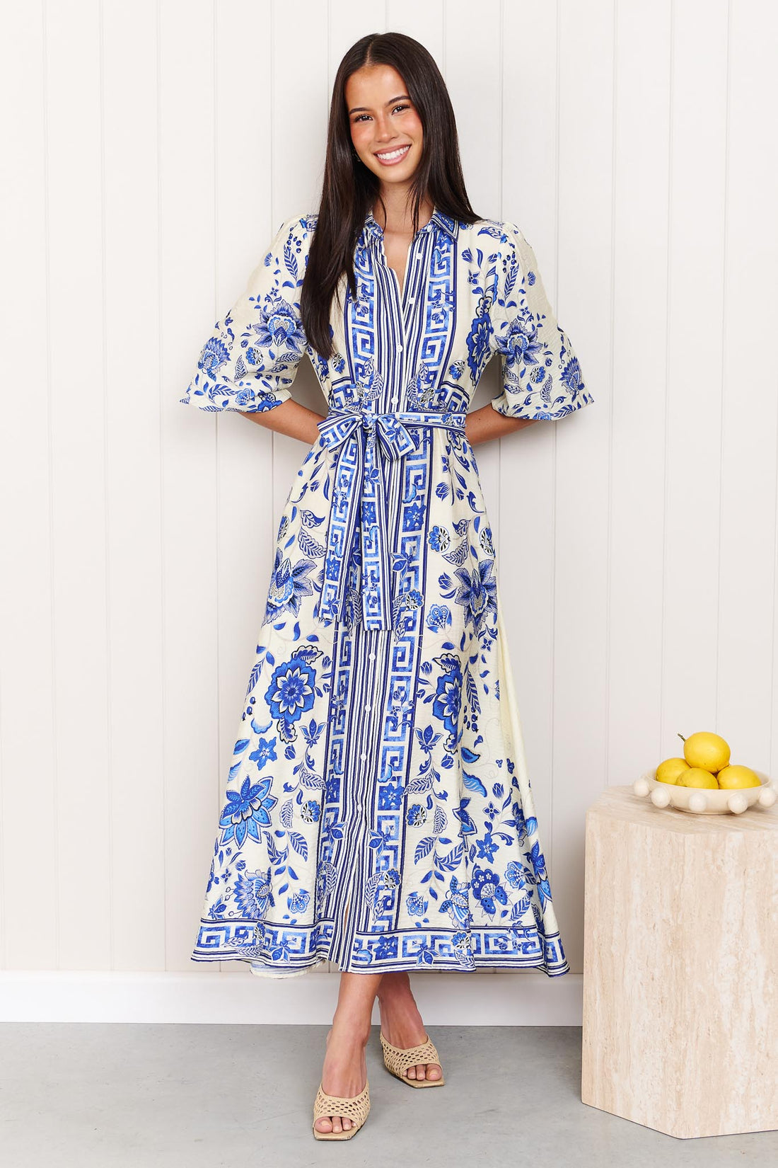 A woman wearing the minnie + MAXXIE Del Mar blue and white floral print midi dress with a tie waist and 3/4 sleeves, standing in front of a white panelled wall.