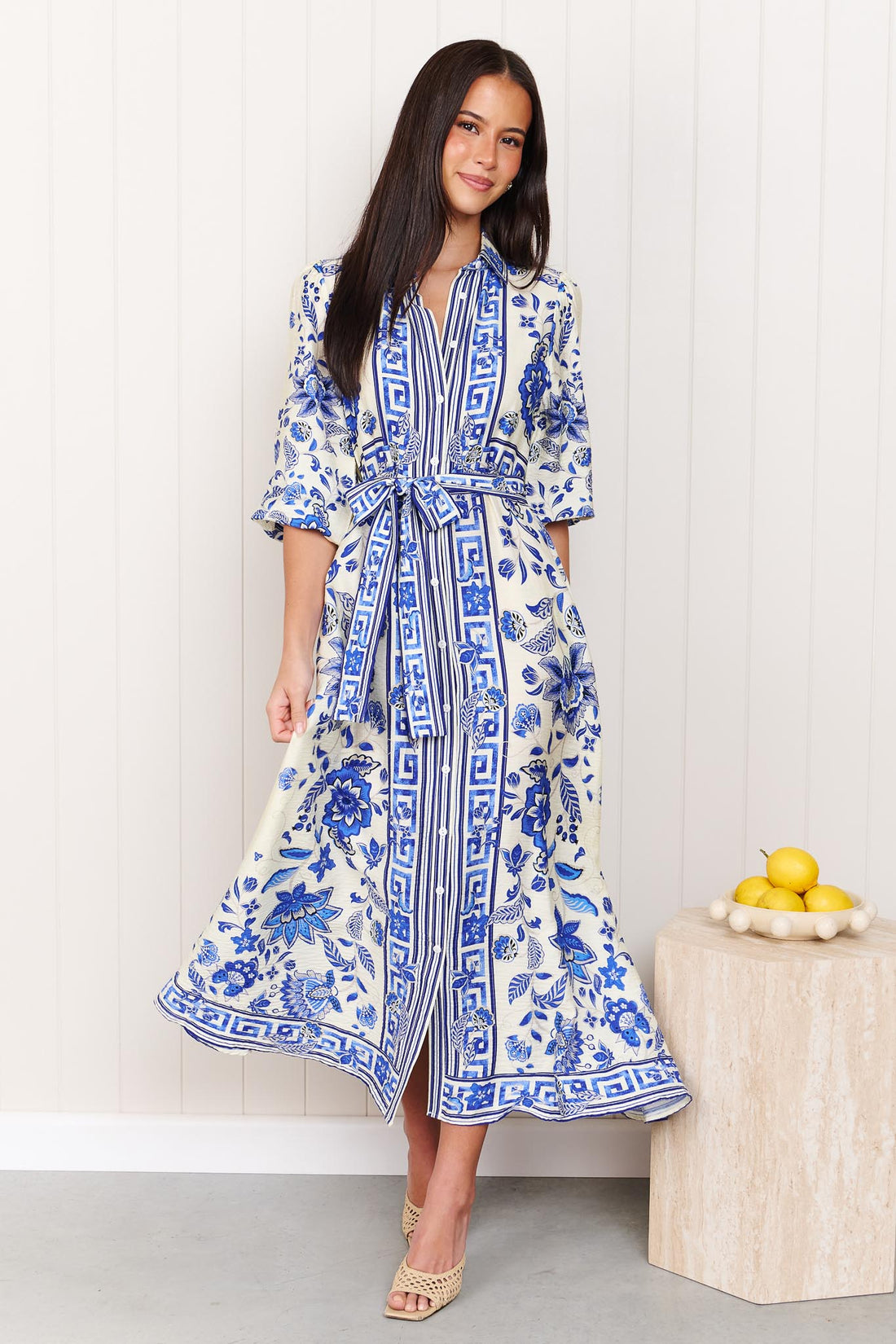 A woman wearing the minnie + MAXXIE Del Mar blue and white floral print midi dress with a tie waist and 3/4 sleeves, standing in front of a white panelled wall.