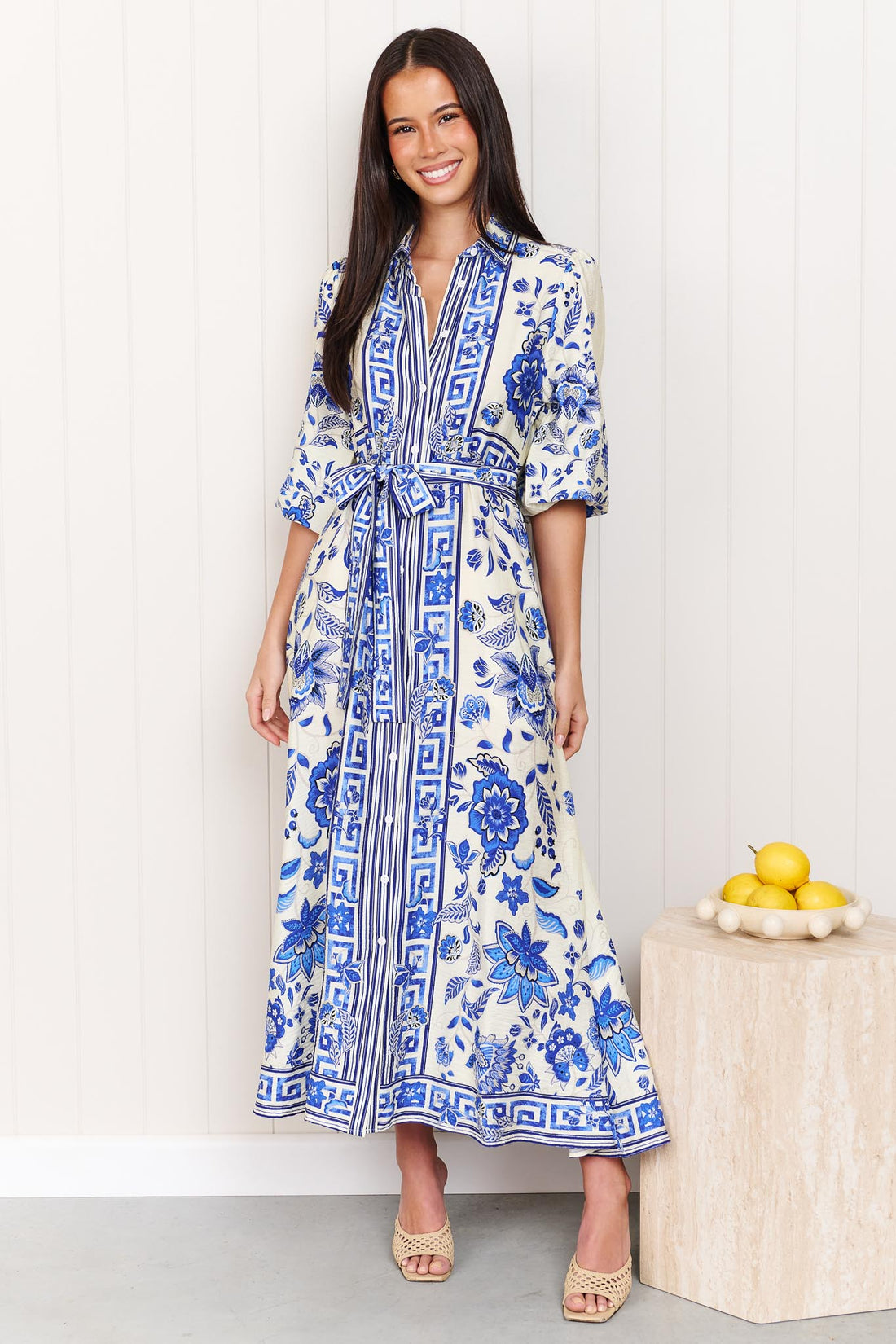 A woman wearing the minnie + MAXXIE Del Mar blue and white floral print midi dress with a tie waist and 3/4 sleeves, standing in front of a white panelled wall.