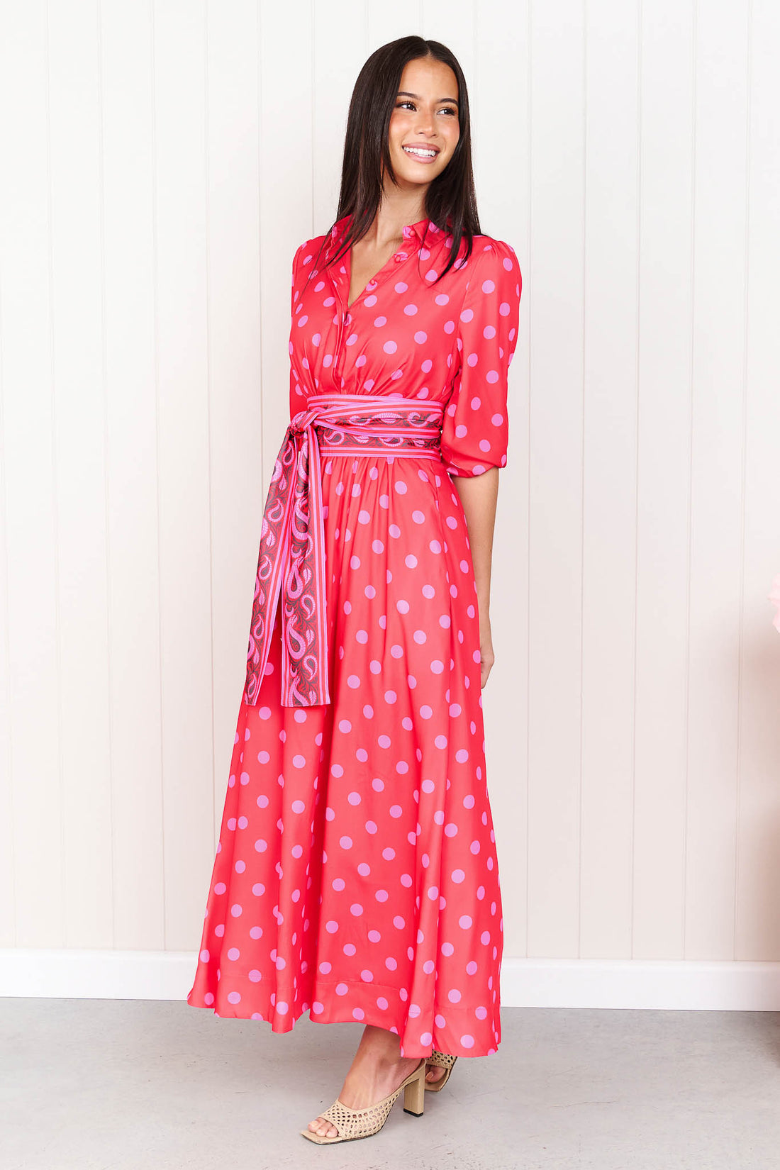 A woman wearing the minnie + MAXXIE Dolce midi event dress in berry with pink polka dots and a matching paisley patterned sash waist tie, standing in front of a white panelled wall.