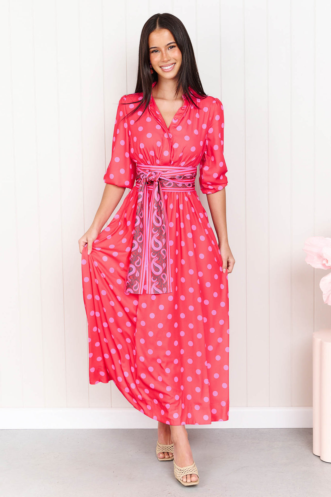 A woman wearing the minnie + MAXXIE Dolce midi event dress in berry with pink polka dots and a matching paisley patterned sash waist tie, standing in front of a white panelled wall.