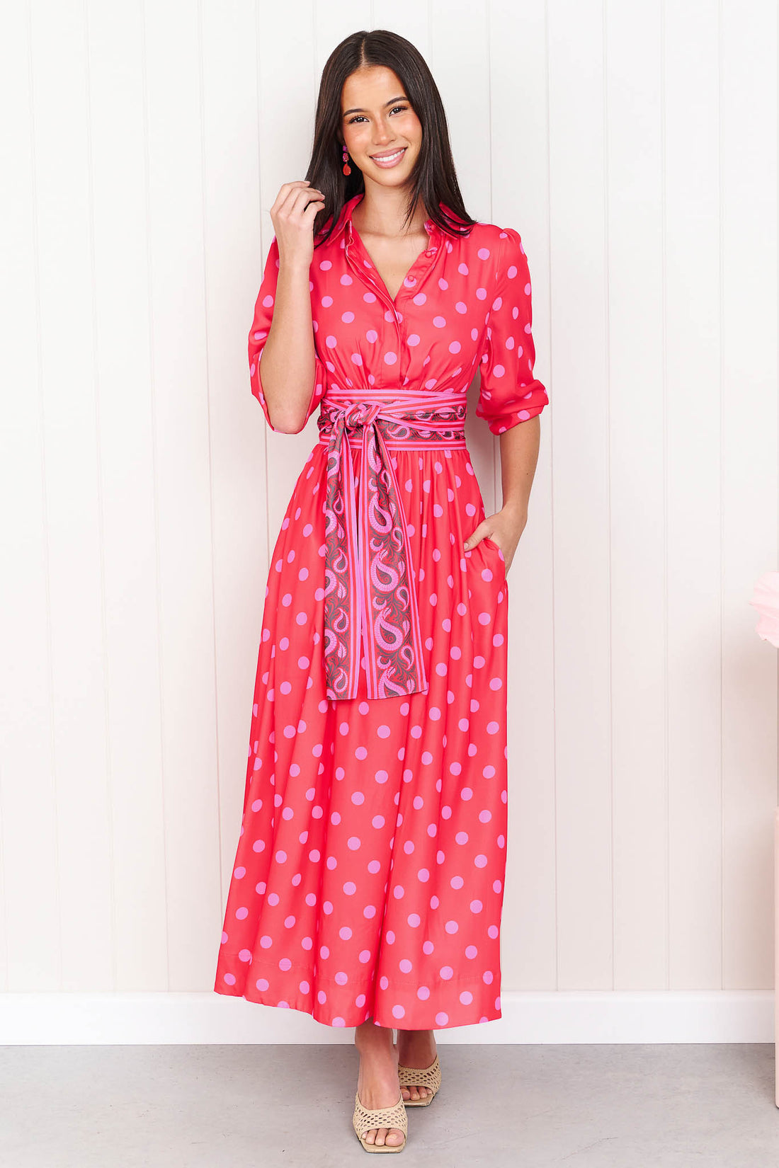 A woman wearing the minnie + MAXXIE Dolce midi event dress in berry with pink polka dots and a matching paisley patterned sash waist tie, standing in front of a white panelled wall.