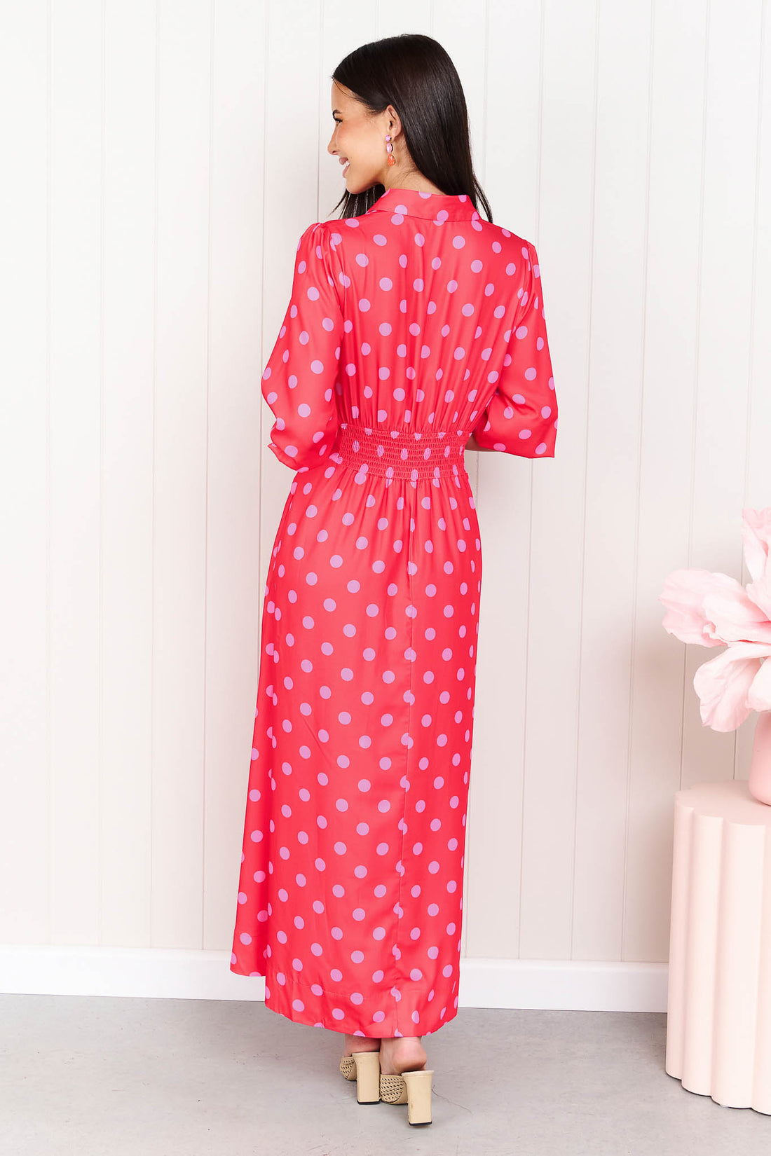 Back view of a woman wearing the minnie + MAXXIE Dolce midi event dress with pink polka dots, standing in front of a white panelled wall.