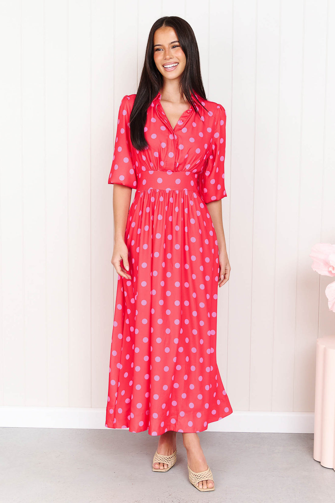 A woman wearing the minnie + MAXXIE Dolce midi event dress with pink polka dots, standing in front of a white panelled wall.