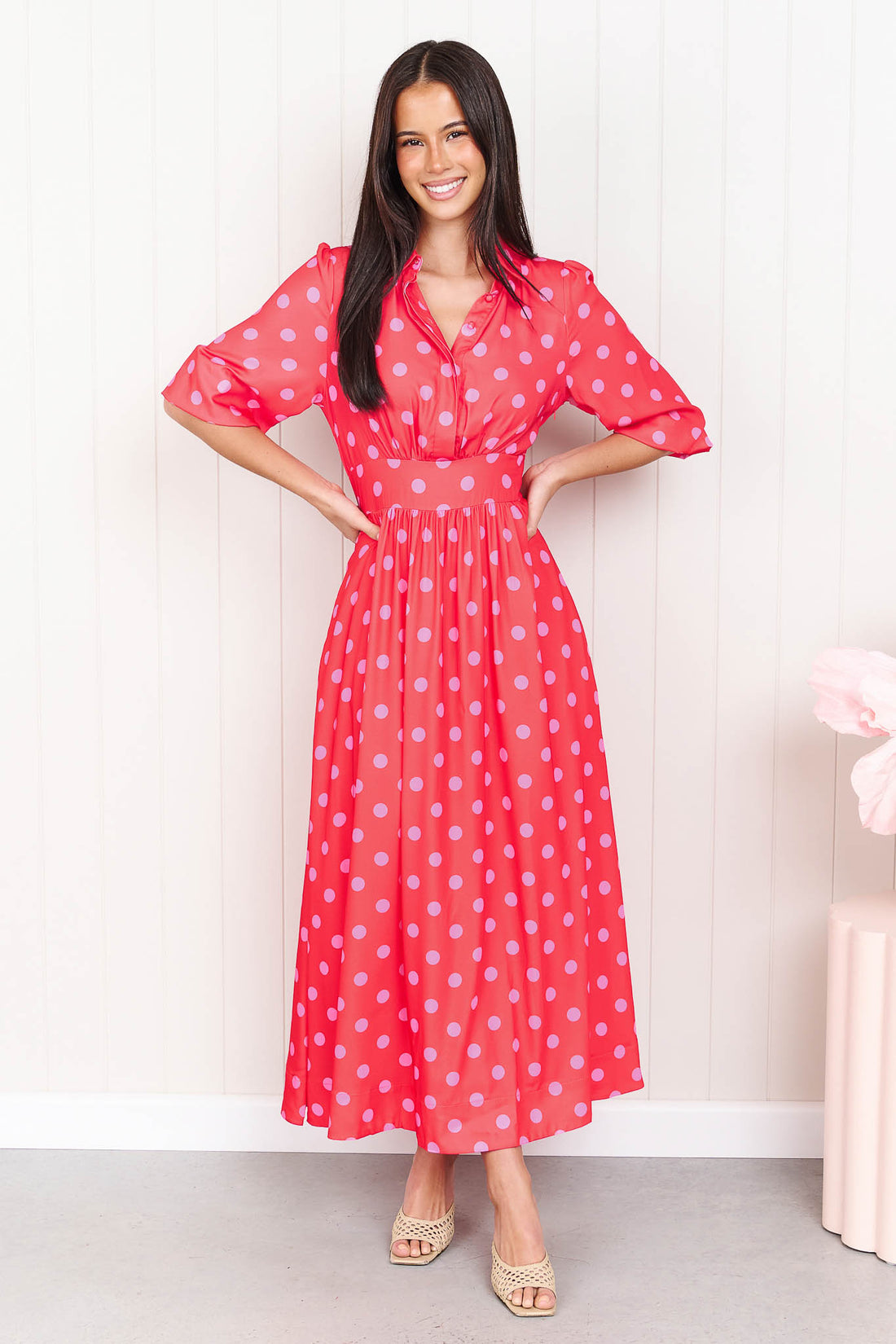 A woman wearing the minnie + MAXXIE Dolce midi event dress with pink polka dots, standing in front of a white panelled wall.
