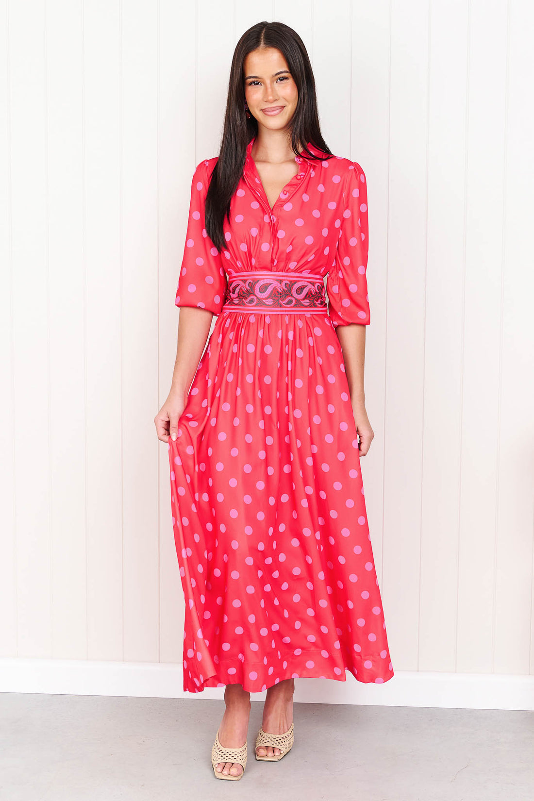 A woman wearing the minnie + MAXXIE Dolce midi event dress with pink polka dots and a matching paisley patterned sash waist tie, standing in front of a white panelled wall.