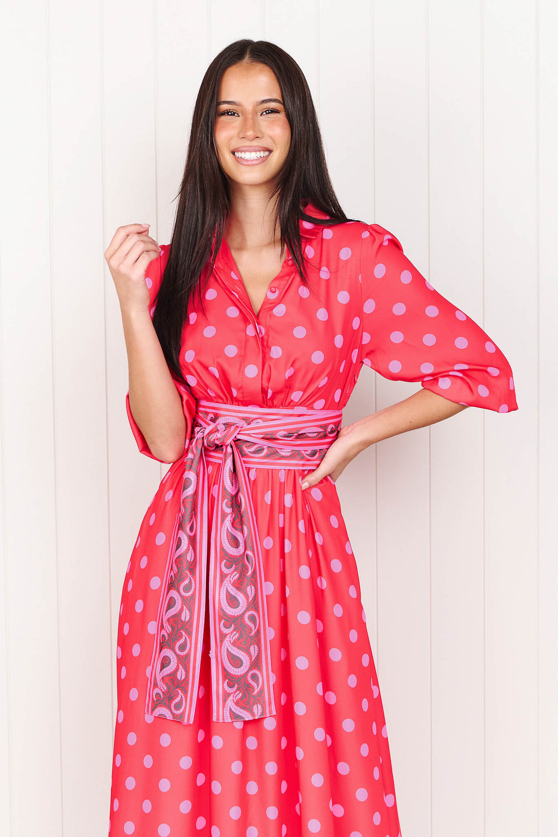 A woman wearing the minnie + MAXXIE Dolce midi event dress in berry with pink polka dots and a matching paisley patterned sash waist tie, standing in front of a white panelled wall.