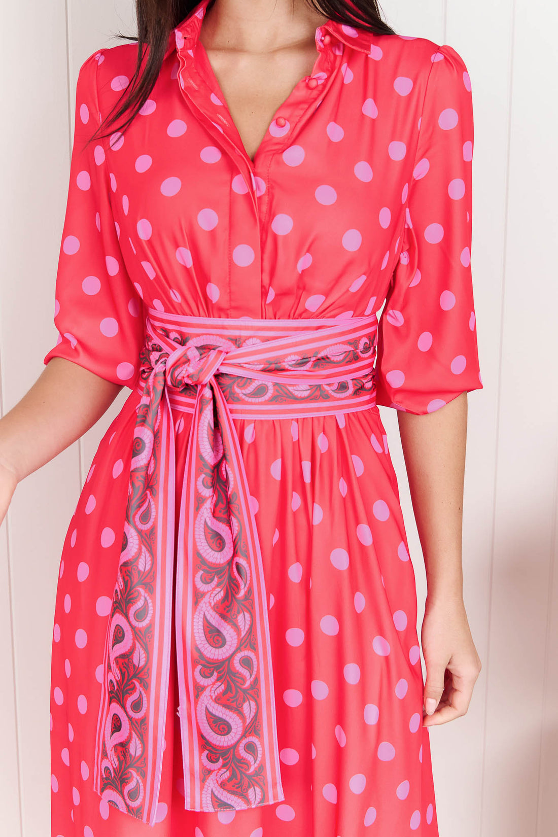 Detail of a woman wearing the minnie + MAXXIE Dolce midi event dress in berry with pink polka dots and a matching paisley patterned sash waist tie.