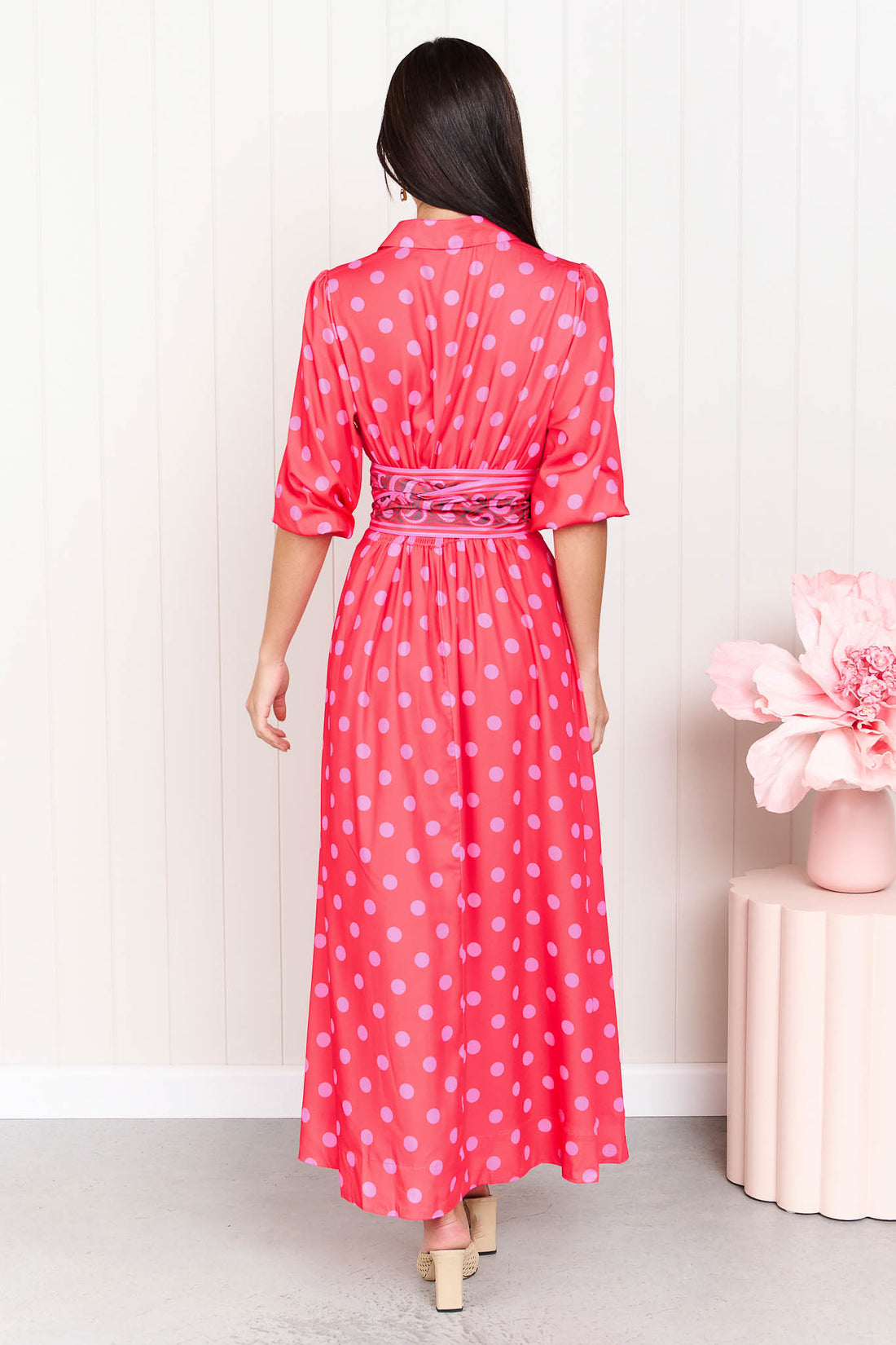 Back view of a woman wearing the minnie + MAXXIE Dolce midi event dress in berry with pink polka dots and a matching paisley patterned sash waist tie, standing in front of a white panelled wall.
