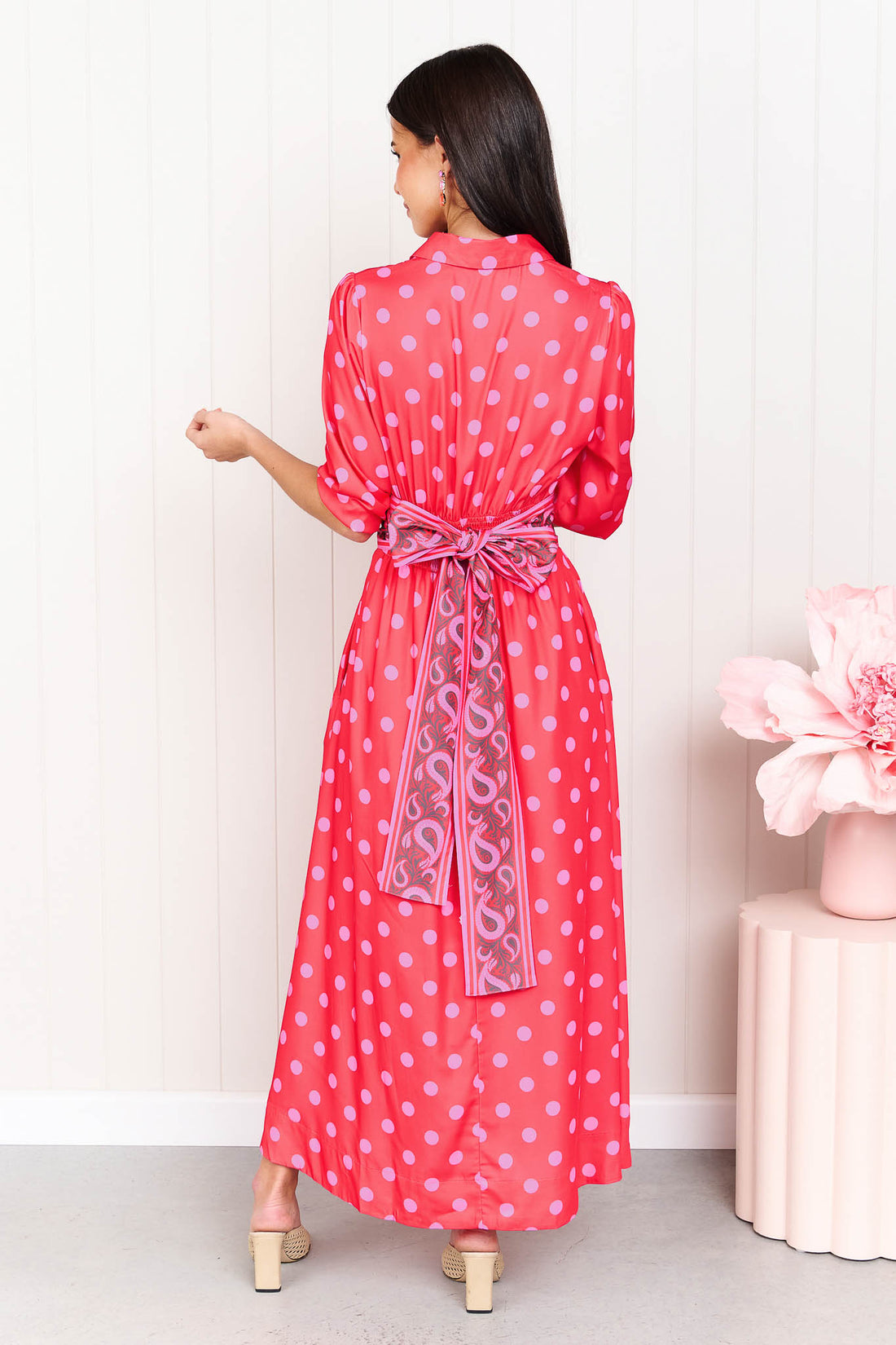 Back view of a woman wearing the minnie + MAXXIE Dolce midi event dress with pink polka dots and a matching paisley patterned sash waist tie, standing in front of a white panelled wall.