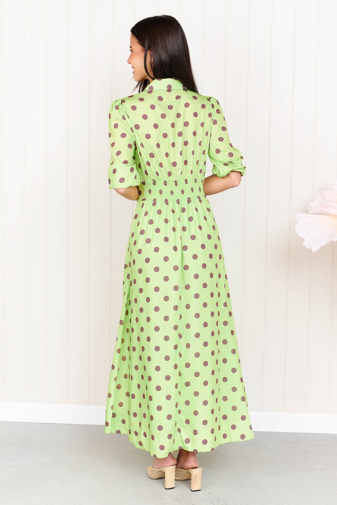 Back view of a woman wearing the minnie + MAXXIE Dolce Midi event dress in lime with chocolate brown polka dots, standing in front of a white panelled wall.