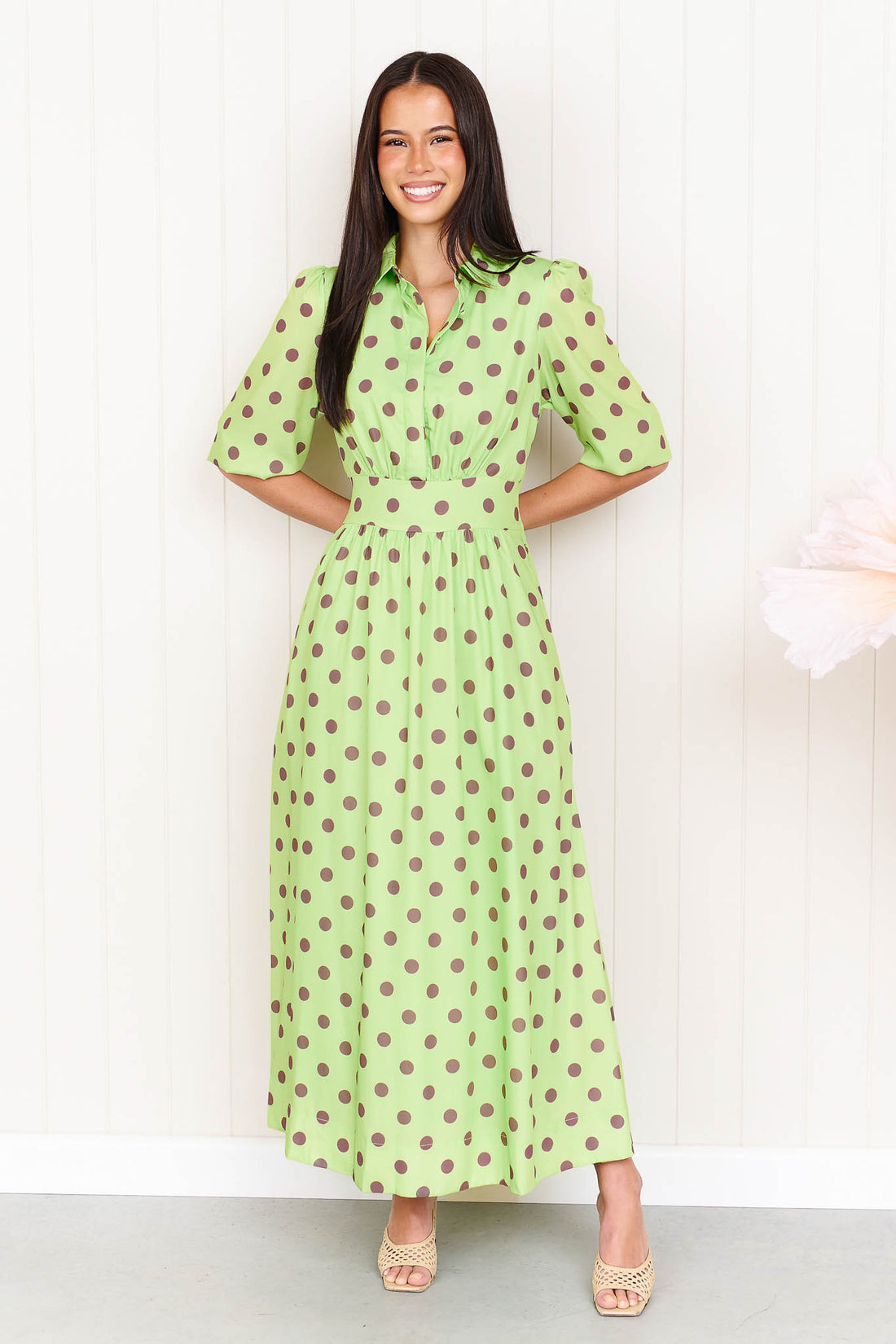 A woman wearing the minnie + MAXXIE Dolce Midi event dress in lime with chocolate brown polka dots, standing in front of a white panelled wall.